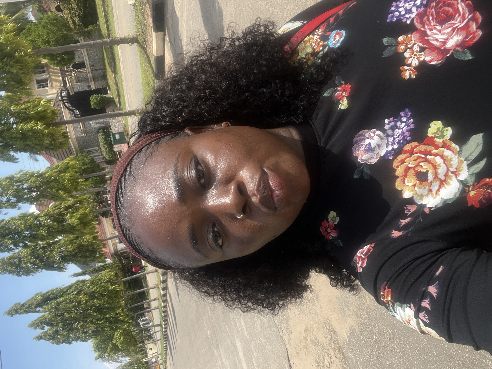 A woman of African descent is depicted in a close-up outdoor selfie, facing the camera with slightly pursed lips. She has dark skin, dark curly hair partially held back by a brown headband, and a silver hoop nose ring in her right nostril. Small gold hoop earrings are visible. She is wearing a black top adorned with a vibrant floral print featuring large red, orange, purple, and white flowers with green leaves. The background shows an urban street scene under clear skies, with an asphalt road and a black and white striped curb. A consistent line of tall, slender, conical green trees (likely *Polyalthia longifolia*) borders the road, extending into the distance. Beyond the trees, residential buildings with distinctive tiled roofs and ornate gates are visible. A person in a red shirt is faintly discernible walking in the far background. The architectural style, street planning, and prevalence of specific tree species are characteristic of developed areas within Abuja, Nigeria.