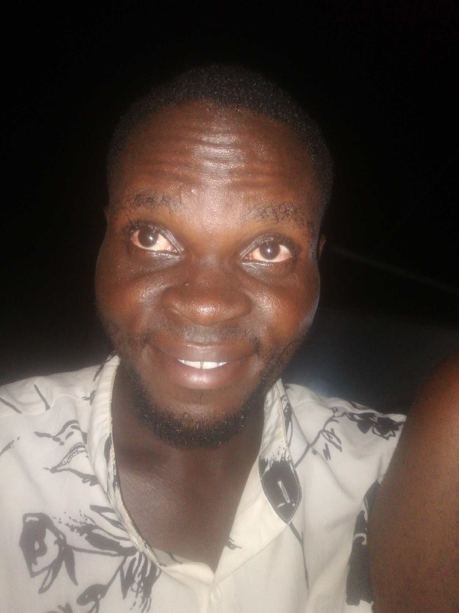 A close-up, front-facing portrait of a dark-skinned man with short, dark hair, a beard, and a slight smile. He is wearing a light-colored shirt with a dark floral pattern. The background is dark, suggesting a nighttime setting. Ikorodu, Nigeria is the location. No other details are discernible.