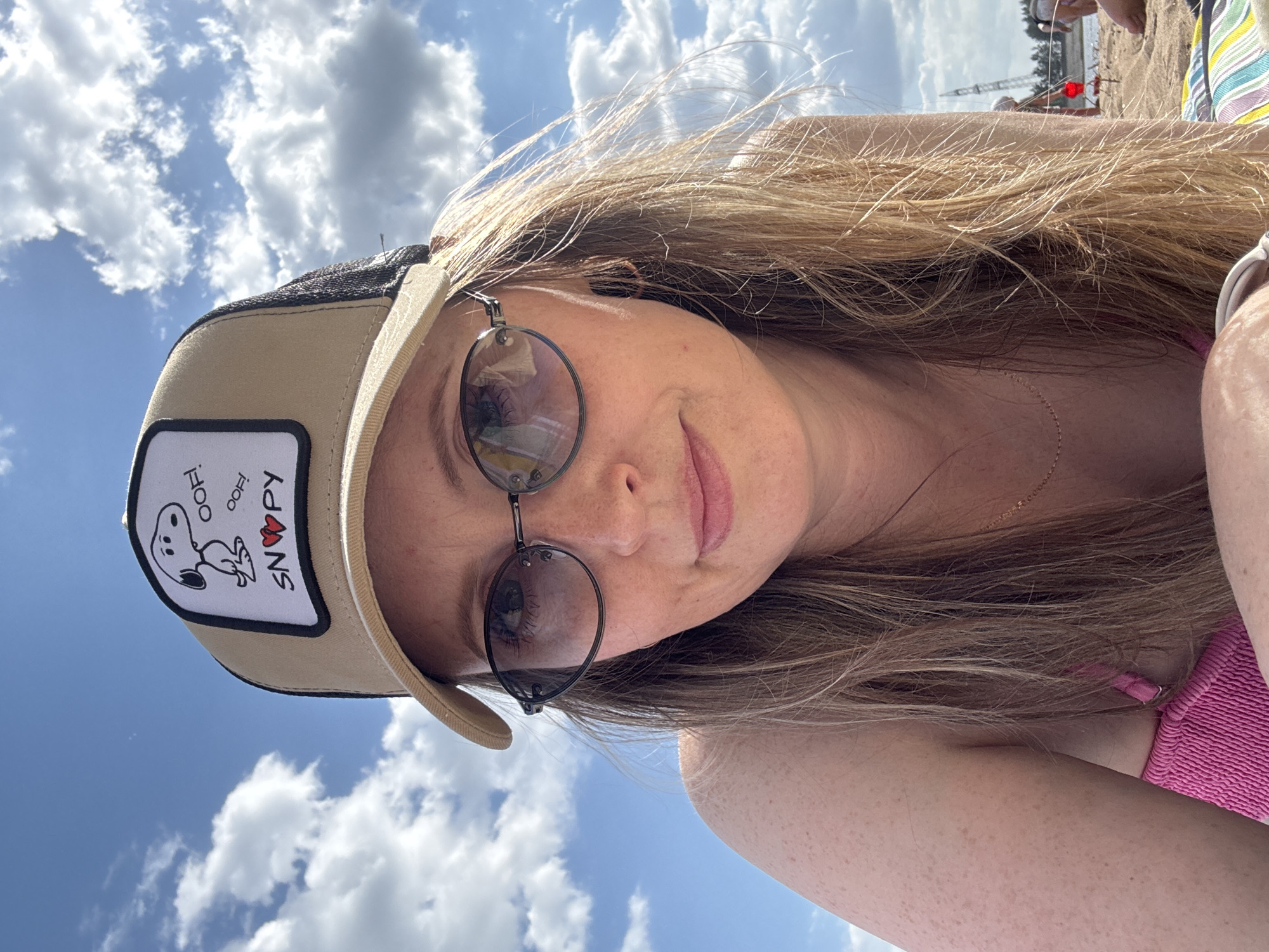A woman with fair skin, light brown hair, and wearing sunglasses and a tan baseball cap with a Snoopy patch is posing for a selfie. She is wearing a pink top. The background shows a blue sky with fluffy white clouds. In the far background, the scene appears to be a beach with people, a tall tower, and possibly a body of water. The location is Chaikovsky, Russia.
