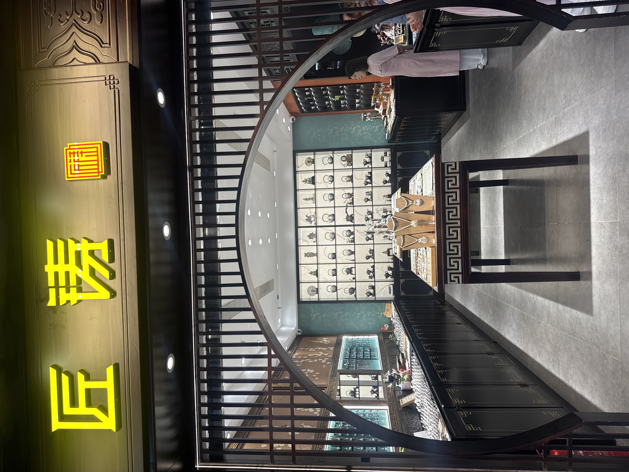 The image depicts the entrance and partial interior of a retail store, presumed to be located in Qingyang District, China. Above the dark wooden entrance facade, a neon yellow-green sign displays three Chinese characters: "匠" (jiàng, craftsman), "铸" (zhù, to cast/forge), and a stylized square character resembling a seal script "坊" (fāng, workshop/store), together reading "匠铸坊" (Craftsman's Forge/Casting Workshop).
The entrance features a dark wooden archway framed by vertical wooden slats above. Inside, the store utilizes a dark wood aesthetic with intricate patterns. On the left interior wall, a brown panel displays white floral patterns, resembling cherry blossoms. The right interior wall is covered in a teal-colored wallpaper with subtle light patterns. The ceiling features multiple recessed spotlights providing bright illumination.
In the center of the foreground, a dark wooden display table with white Greek key patterns on its side supports multiple jewelry busts showcasing ornate silver-toned necklaces. Along the left side of the store, a long, dark wooden counter with cabinets underneath is topped with numerous small, shiny silver items, possibly rings or bracelets. Further back on the left, additional illuminated display cases are visible.
On the right side of the store, a large wall-mounted display unit with square compartments showcases a collection of silver-toned metalware, predominantly small teapots or kettles, alongside a few figurines including a horse and rider, and a Buddha statue. Adjacent to this, a narrower vertical display holds multiple small white cups. A woman in a light pink tunic dress stands to the right, facing towards the left side of the store. Her hands are clasped in front of her. Partially visible on the far right are other individuals in light-colored clothing. The floor is covered in light gray tiles.