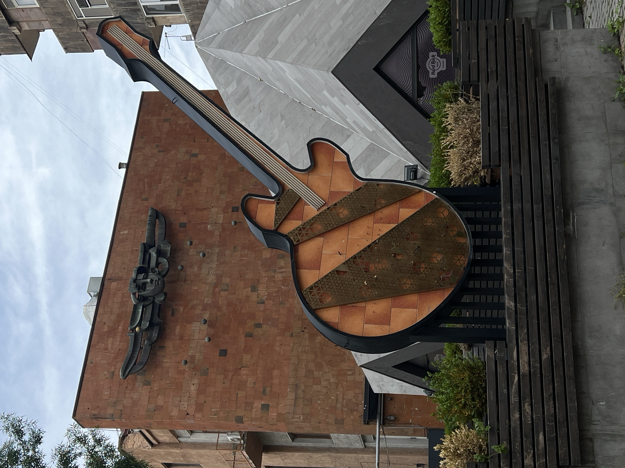 The image shows an outdoor scene in Unknown, Unknown, featuring a large, stylized guitar sign attached to the facade of a building. The guitar is predominantly tan with brown stripes and a black outline. The guitar's neck extends upward towards a cloudy sky. To the left of the guitar, on the brick building, is a dark, abstract sculpture. The building adjacent to the brick structure has a gray and white facade with angular architectural features. A Hard Rock Cafe logo is partially visible in a window of this building. Wooden planks and greenery are present to the right of the guitar. The scene appears to be a street view, with other buildings partially visible in the background.