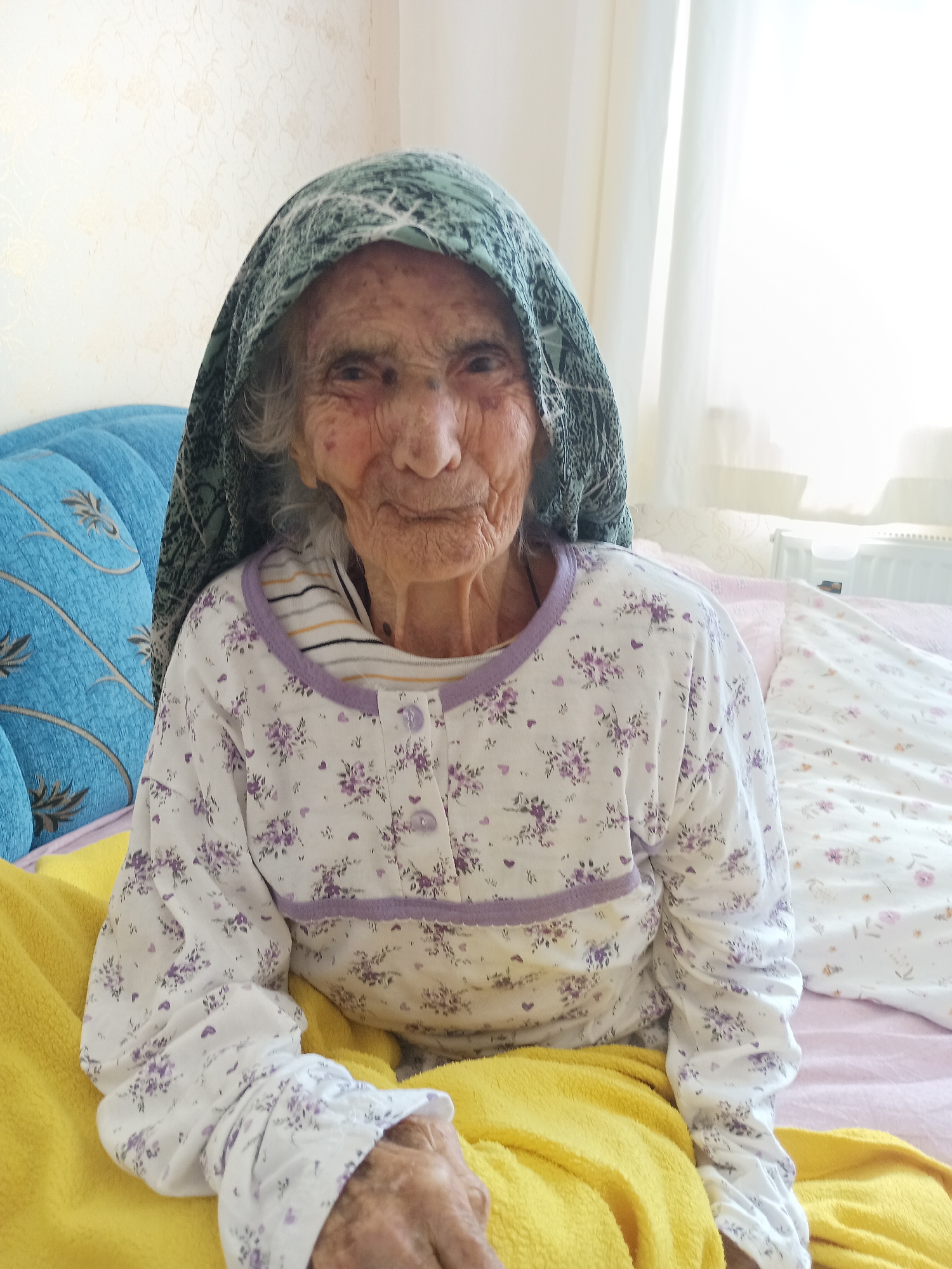 The image shows an elderly woman wearing a headscarf and a floral print pajama top. She is lying in bed, with a blue upholstered headboard on her left and a white, floral patterned duvet or bedspread on her right. A yellow blanket is draped over her. The background appears to be an indoor setting, possibly a bedroom, with a patterned wallpaper and curtains visible. The light suggests it is daytime, possibly near a window. The image has been taken from a medium distance, focusing on the woman's upper body and face. The location is identified as Bahşılı Belediyesi, Turkey.