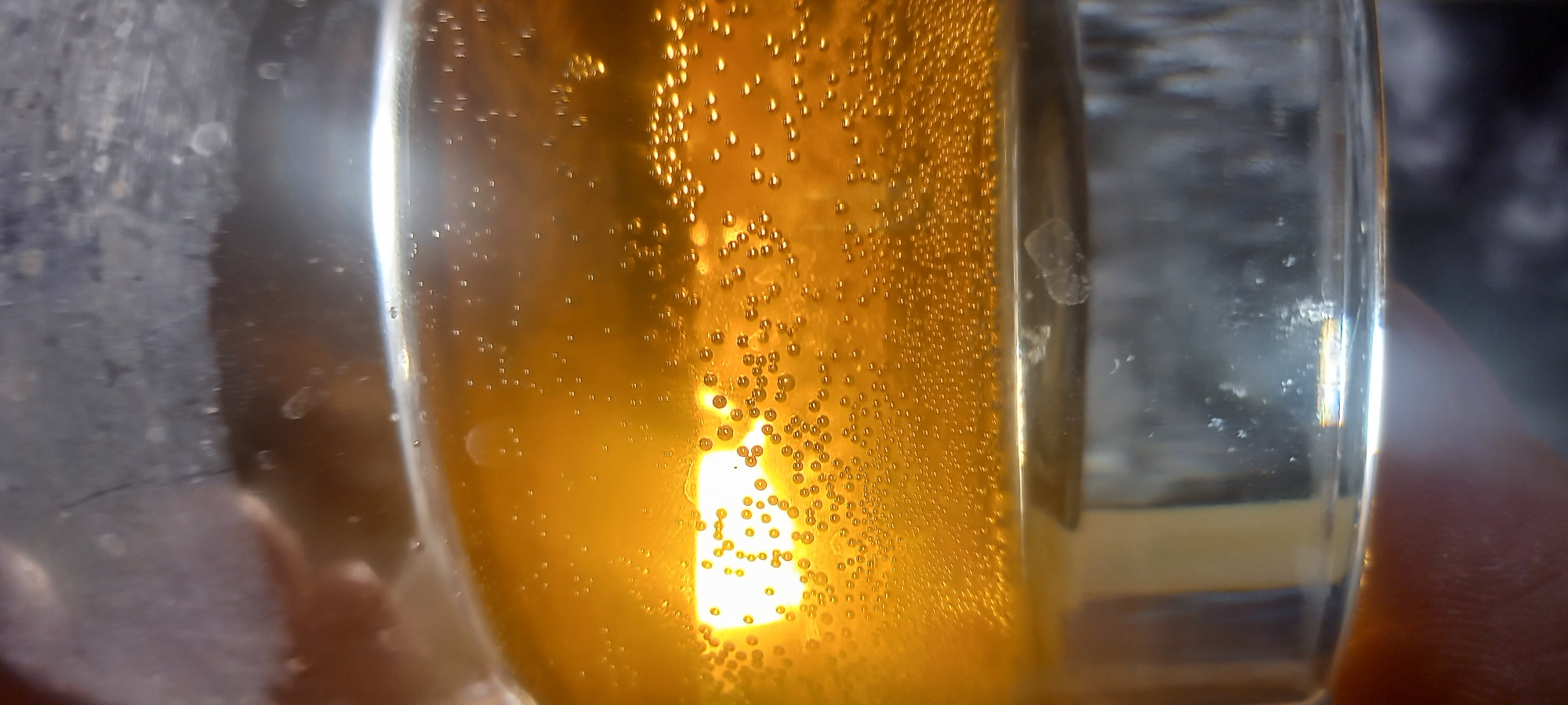 A close-up shot reveals a glass of golden-hued liquid filled with numerous small bubbles. The glass itself is clear, with some minor smudges and imperfections visible on the surface. Light refracts through the liquid, creating a bright spot near the center. The image does not depict a location in Kryvyi Rih, Ukraine or any people or actions. It solely shows a close-up view of the liquid and glass.