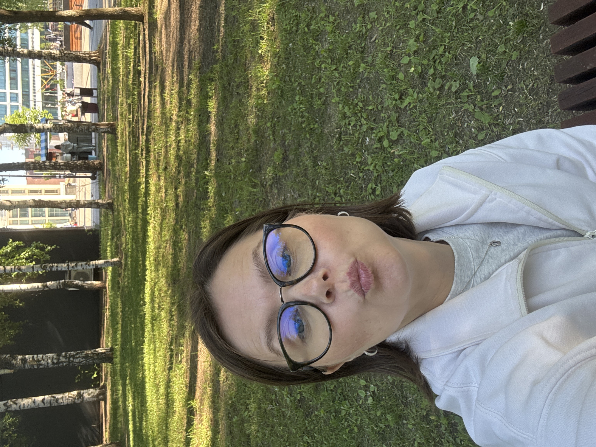 A young woman with brown hair, wearing glasses and a white jacket over a gray sweatshirt, poses for a selfie in a park. She is making a kissing face. The background shows a grassy area with trees and in the distance, buildings and people. The scene is set in Izhevsk, Russia, likely in a public park like Udmurt Republic National Library Park or Park of Kirova, given the presence of birch trees and a modern urban backdrop. The lighting suggests it is daytime, possibly late afternoon due to the warm sunlight casting shadows.