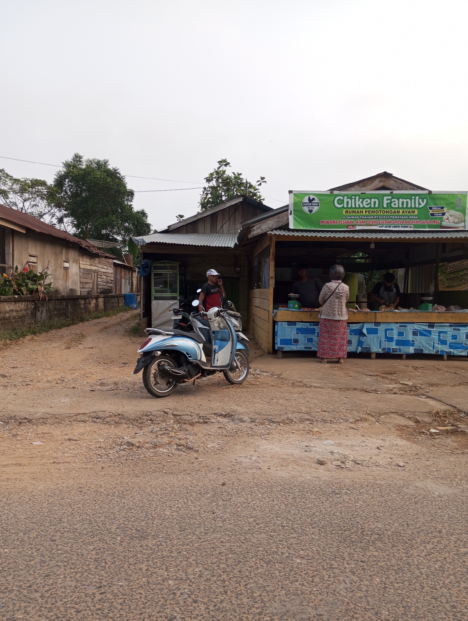 The image depicts a street scene in Pematang Reba, Indonesia. On the right, a small open-air market stall with a green banner advertising "Chiken Family RUMAH PEMOTONGAN AYAM" (Chicken Family Chicken Slaughterhouse) is visible. The banner also lists the address "JL.AHMAD THAHAR RT.02/010 PEMATANG REBA". Two men are working behind the counter of the stall, and a woman wearing a patterned skirt and a beige jacket is standing facing the counter. To the left of the stall, a blue and white scooter is parked. Further to the left, a dirt road leads past a weathered building. The sky is overcast.