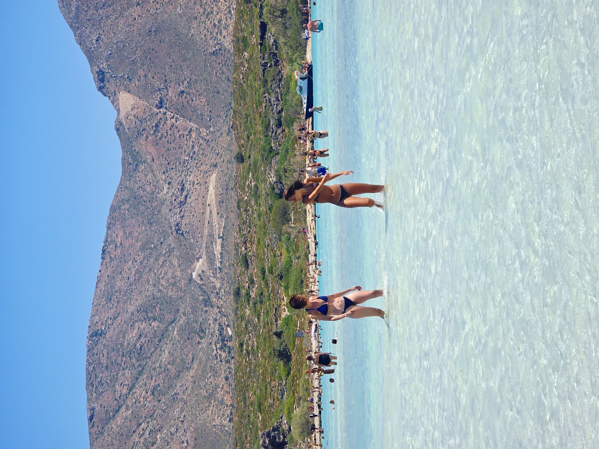Two young women in bikinis are wading in the shallow, crystal-clear turquoise water of a beach. The woman on the left, wearing a blue bikini, stands facing towards the right. The woman on the right, in a black bikini, is facing left, with her arms outstretched as if in motion. Both women appear to be enjoying the water. In the background, the beach is populated with numerous other people, some swimming, others standing and conversing. A white boat with a dark stripe is anchored in the water. The scene is set against a backdrop of arid, rocky hills dotted with green vegetation, under a clear blue sky. The overall environment suggests a sunny, tropical or Mediterranean beach destination.