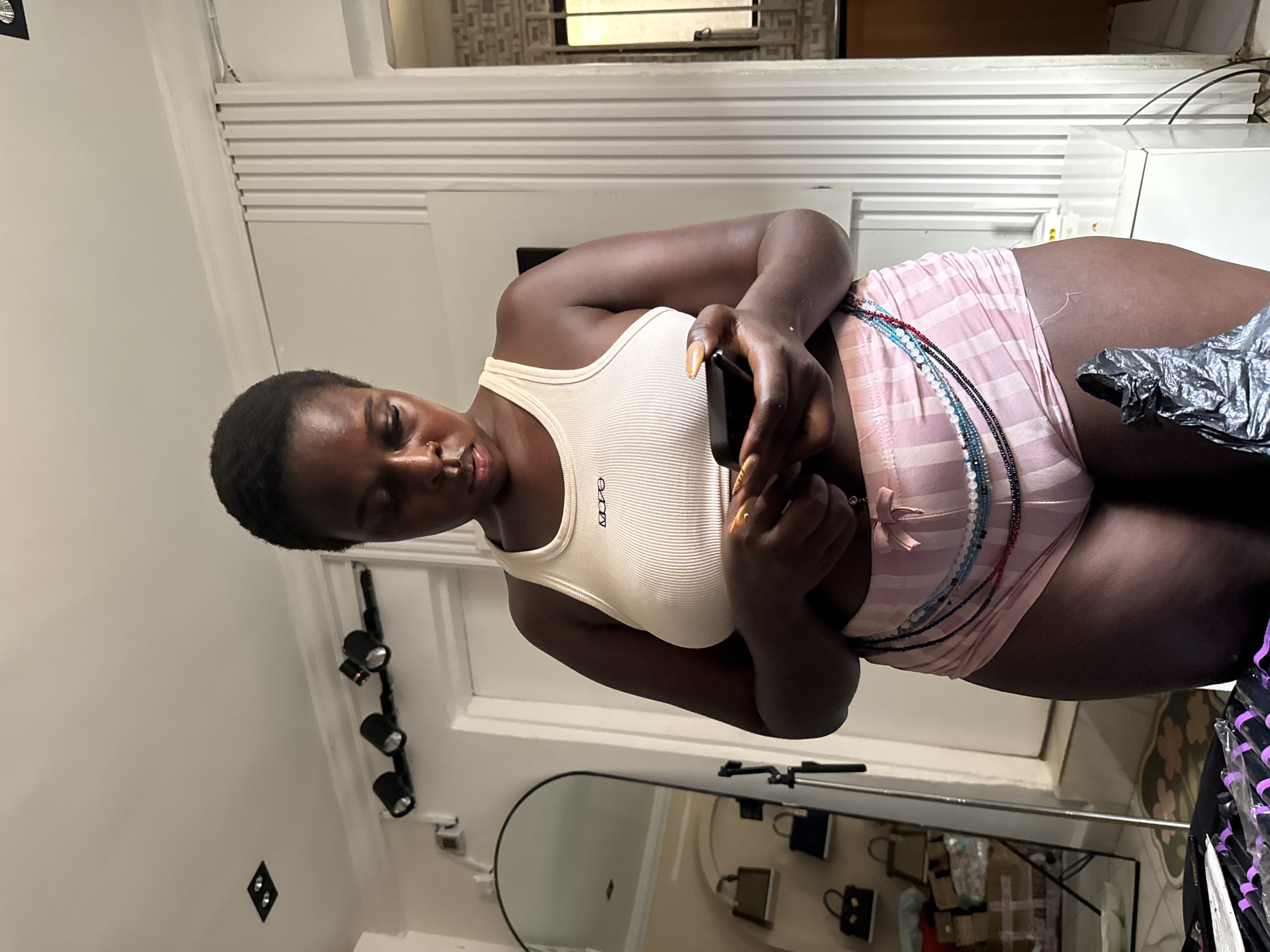 A dark-skinned woman with short hair is standing and looking down at her phone. She is wearing a white ribbed tank top and pink striped shorts. Around her waist, she has a beaded necklace. The woman is indoors, in what appears to be a room with a mirror on the left side of the frame. The background includes a door, a wall with vertical slats, and a window with a patterned grate. There are also some bags visible on shelves in the mirror's reflection. The lighting suggests it is daytime. The location is identified as Abuja, Nigeria.