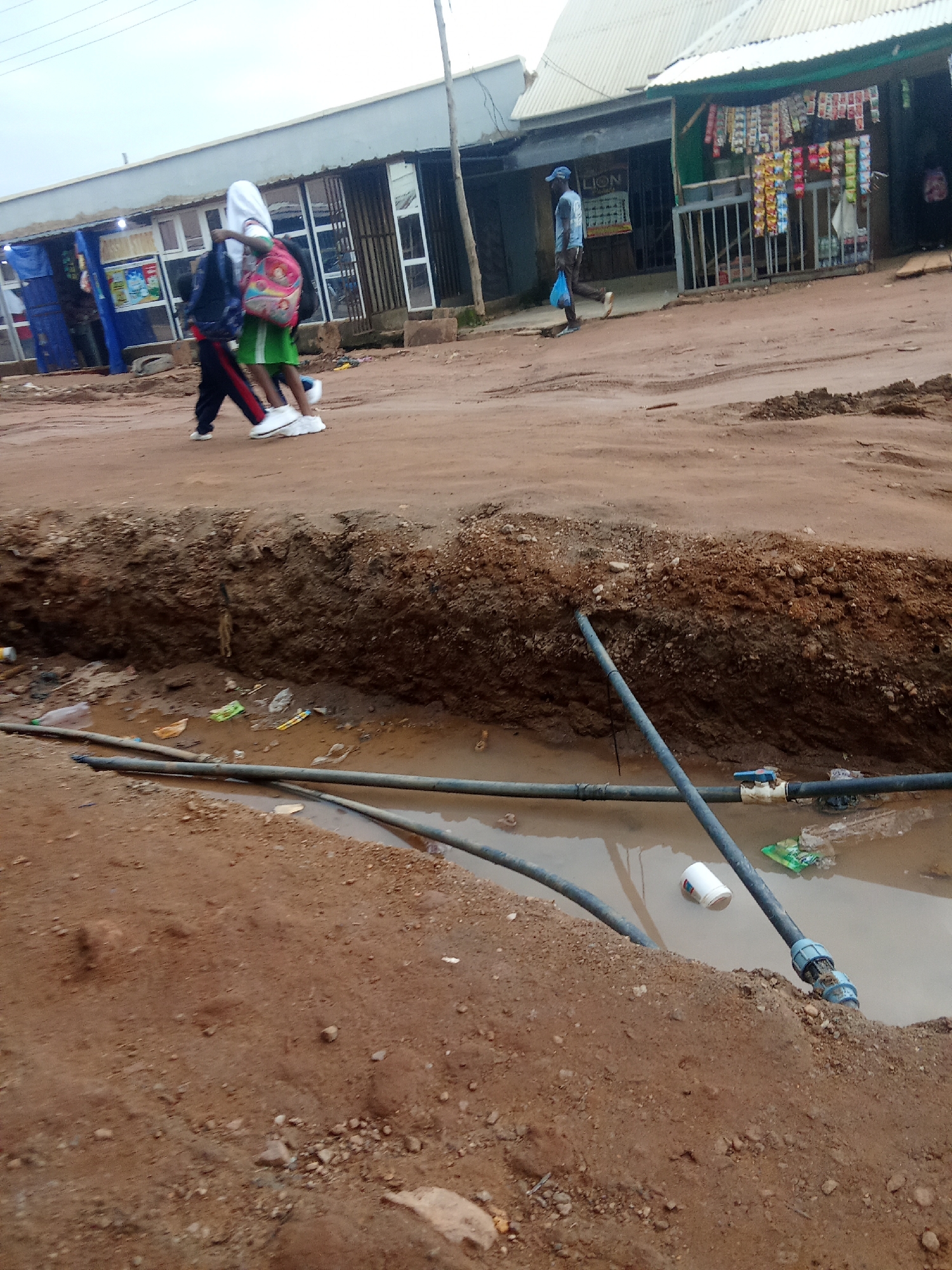 In Kagini, Nigeria, a dirt road is under construction, with a deep trench dug into the side of the road. Several pipes run along the bottom of the trench, which is filled with murky water. Debris, including plastic bottles and bags, is scattered in the water. Two young people, one wearing a white hijab and the other a green skirt, walk across the road carrying backpacks. Another person in a blue cap and shirt walks in the background, carrying a blue plastic bag. Shops with metal shutters line the street, displaying various goods for sale. The sky is overcast, suggesting a cloudy day.