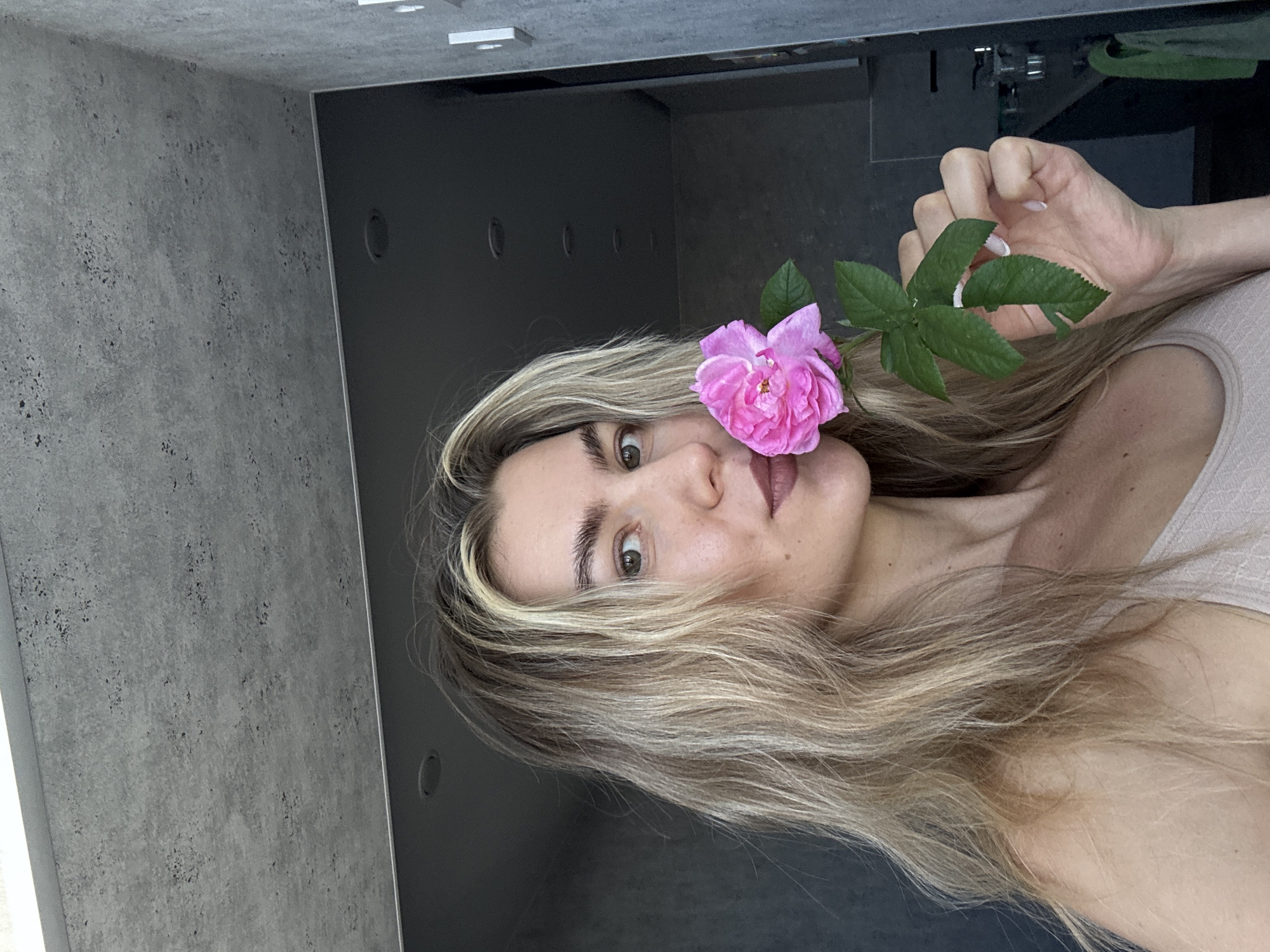 The image displays a fair-skinned woman with long, wavy blonde hair with lighter highlights. Her hair cascades over her shoulders and partially frames her face. She appears to be in her 20s or 30s, with light-colored eyes looking directly at the viewer and defined eyebrows. Her lips are adorned with a muted, reddish-brown color. She is wearing a light-colored, possibly beige or cream, garment, only a small portion of which is visible on her upper body.
She holds a vibrant pink flower with multiple green leaves close to her face, positioned to the left of her nose, partially covering her cheek. Her right hand, featuring light-colored fingernails, is visible gripping the stem of the flower.
The setting appears to be indoors, suggesting a modern or contemporary space. To the left of the woman, a grey, textured wall, possibly concrete or a similar industrial finish, occupies a significant portion of the background. Above her head and to her right, dark, smooth surfaces, which could be cabinets, a door, or wall panels, are visible. Some recessed lights are faintly discernible on what appears to be a ceiling in the upper part of the frame. The lighting is soft and ambient, without strong shadows.
The woman maintains a direct gaze towards the camera, conveying a serene and focused expression. The image is captured with a vertical orientation, causing the woman's head and body to appear tilted 90 degrees to the right relative to a standard upright posture. There is no visible text in the media file.