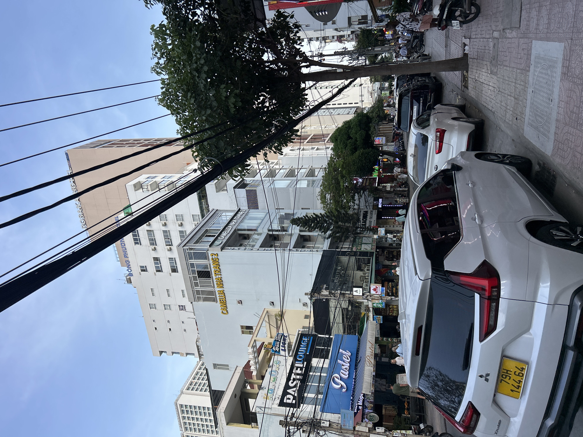 The image shows a street scene in Nha Trang Ward, Vietnam. A white Mitsubishi Xpander SUV is parked on the right side of the street, its license plate reads "79H 144.64". Behind it, a white Kia sedan and a dark-colored Kia SUV are also parked along the curb. Several buildings line the street, including one with a prominent sign for "CAMELLIA NHA TRANG 2". Overhead, a dense network of electrical wires crisscrosses the sky. A large tree stands on the sidewalk, its branches casting shadows. Several people are visible walking on the sidewalk and in the street. The architecture suggests a commercial and residential area. The overall impression is a bustling urban environment on a bright day, indicated by the clear sky and the lighting.