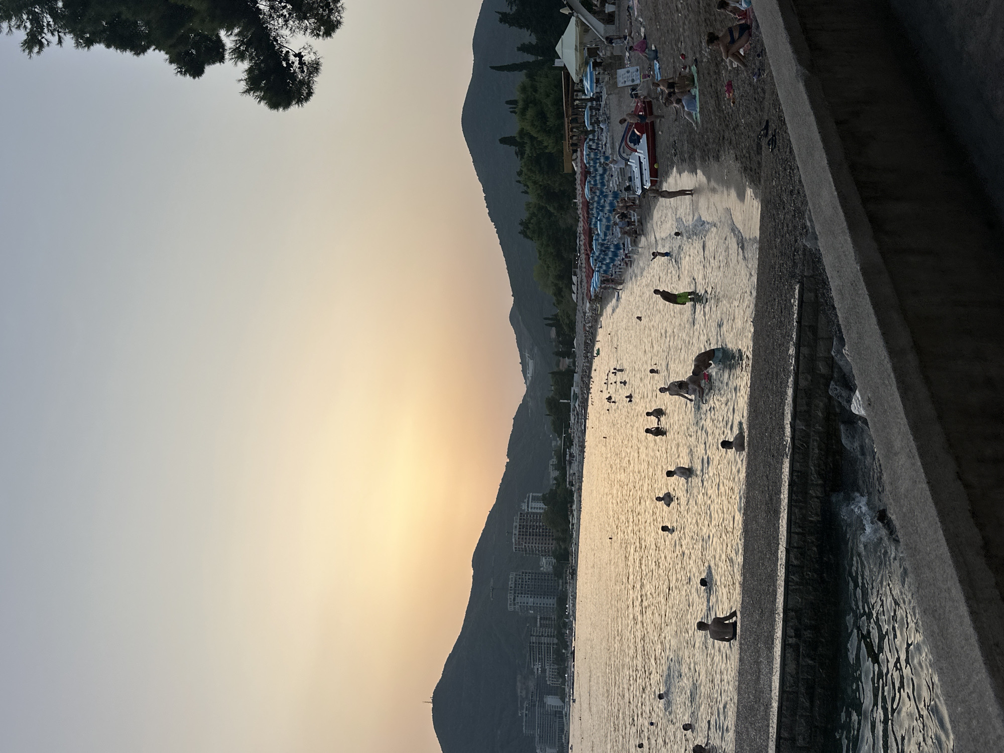 A wide-angle, full-shot image captures a serene beach scene in Budva, Montenegro, during a sunset. The sky transitions from a pale blue at the top to a warm, soft orange glow near the horizon, creating a tranquil ambiance. In the distance, dark, tree-covered mountains form a dramatic backdrop to the scene. Along the coastline, several multi-story buildings and construction sites are visible, hinting at the city's development.
The foreground of the image features a stone embankment overlooking the sea. The water, reflecting the sunset's golden hues, is populated by numerous people swimming, wading, and enjoying the water. Some are standing, while others are partially submerged, their forms silhouetted against the gentle waves. The beach itself, visible to the right of the water, is lined with rows of blue and white striped umbrellas, indicating a popular tourist spot. People are seated on the sand, relaxing, and some are interacting with each other. Several beach chairs and a few pedal boats are also present. A large pine tree branch extends into the upper right corner of the frame, adding a natural element to the composition. The overall atmosphere is one of leisure and relaxation during a beautiful evening by the sea.