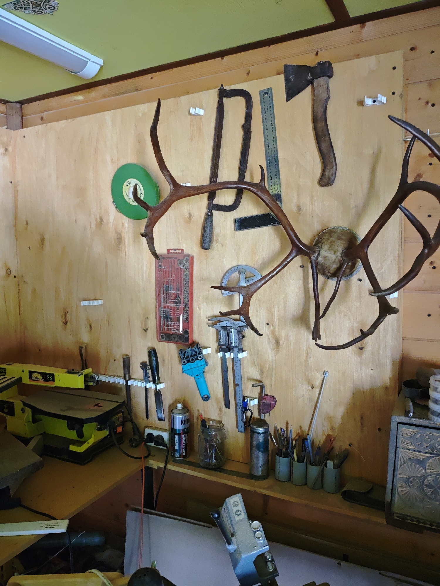 The image shows a cluttered workshop, with a wooden wall covered in tools and various objects. Antlers are mounted prominently on the wall, along with a saw, axe, and ruler. Below the antlers are various tools, including a set of drill bits, a paint can, and containers holding pens and small items. A yellow scroll saw sits on a table in the foreground, and a metal toolbox is visible on the right. The location is Боровково, Russia.