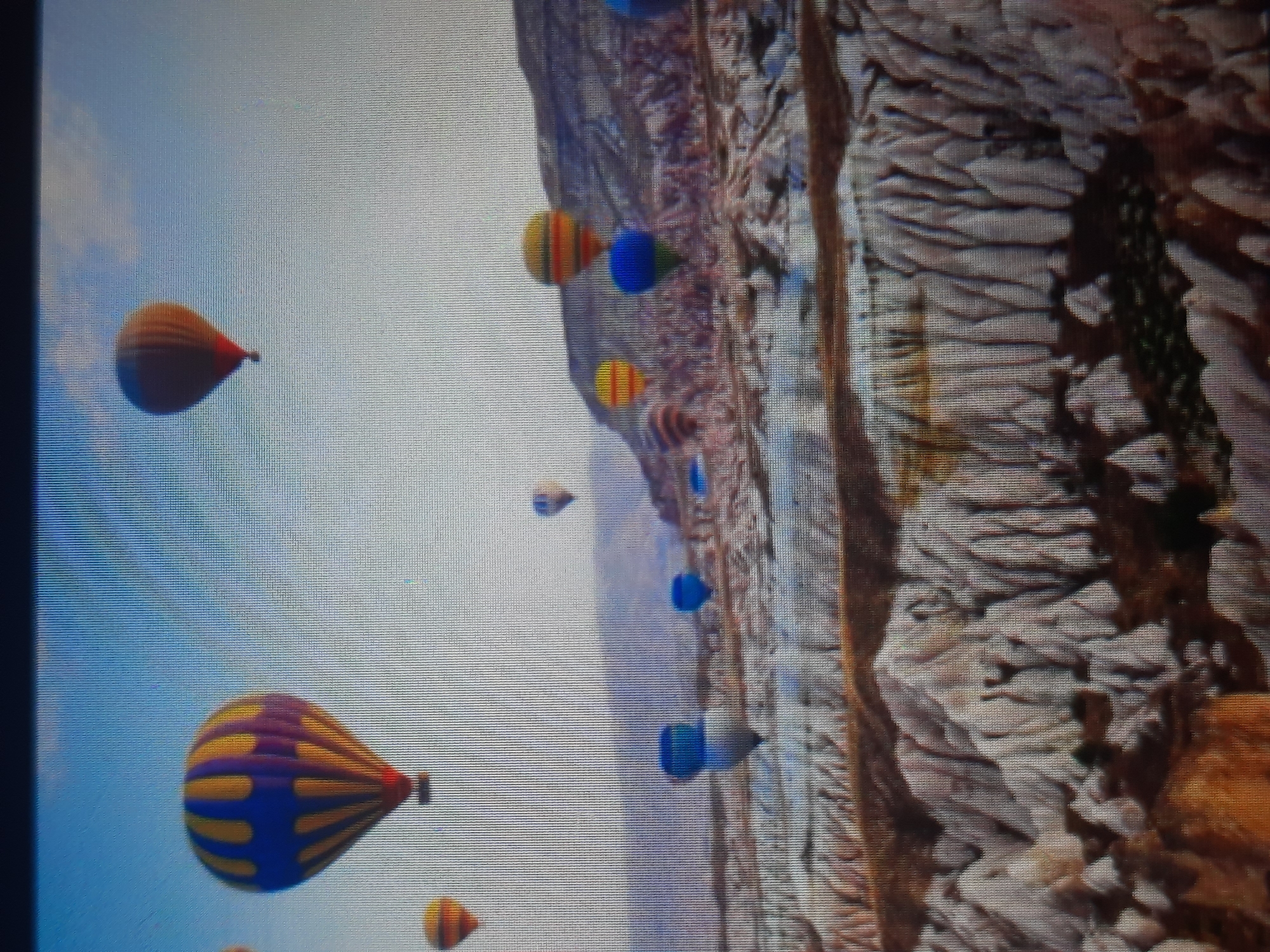 The image depicts a scenic landscape with multiple hot air balloons in the sky. The balloons are of various colors and sizes, with some being closer to the viewer than others. Below the balloons, a unique geological formation of weathered rock formations, resembling canyons or valleys, is visible. The terrain is arid and characterized by sharp, undulating shapes. The sky is clear and blue, suggesting a pleasant day for ballooning. There is no discernible connection to the city of Rosenheim, Germany, as the landscape and atmospheric conditions do not align with typical geographical features of that region. The image focuses solely on the natural environment and the hot air balloons.