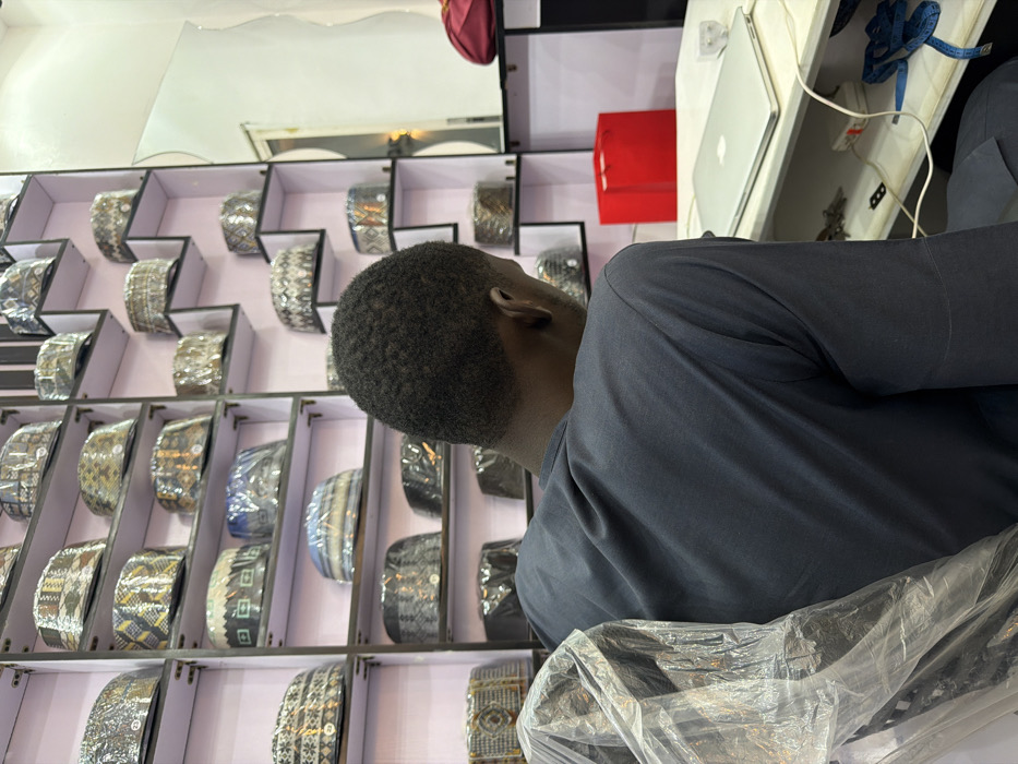 The image displays an indoor scene in Gandu Albasa, Nigeria. An adult individual is seated with their back to the camera, wearing a dark grey long-sleeved garment. Their short, dark, kinky hair is visible. The person is positioned in a chair, partially covered on the left by a clear plastic bag. To the individual's right, a light-colored desk holds a closed silver-colored laptop, a red rectangular box, a white power strip with multiple connected wires, a white charging brick, and a blue coiled measuring tape.
The background consists of a wall lined with numerous shelves, predominantly dark-framed with light backings. These shelves display a large collection of traditional caps, each individually wrapped in clear plastic. The caps exhibit various patterns and colors. On the far right wall, a vertical rectangular mirror is present, reflecting a soft, indistinct light source. Below the mirror, a red fabric item rests on a shelf or surface.