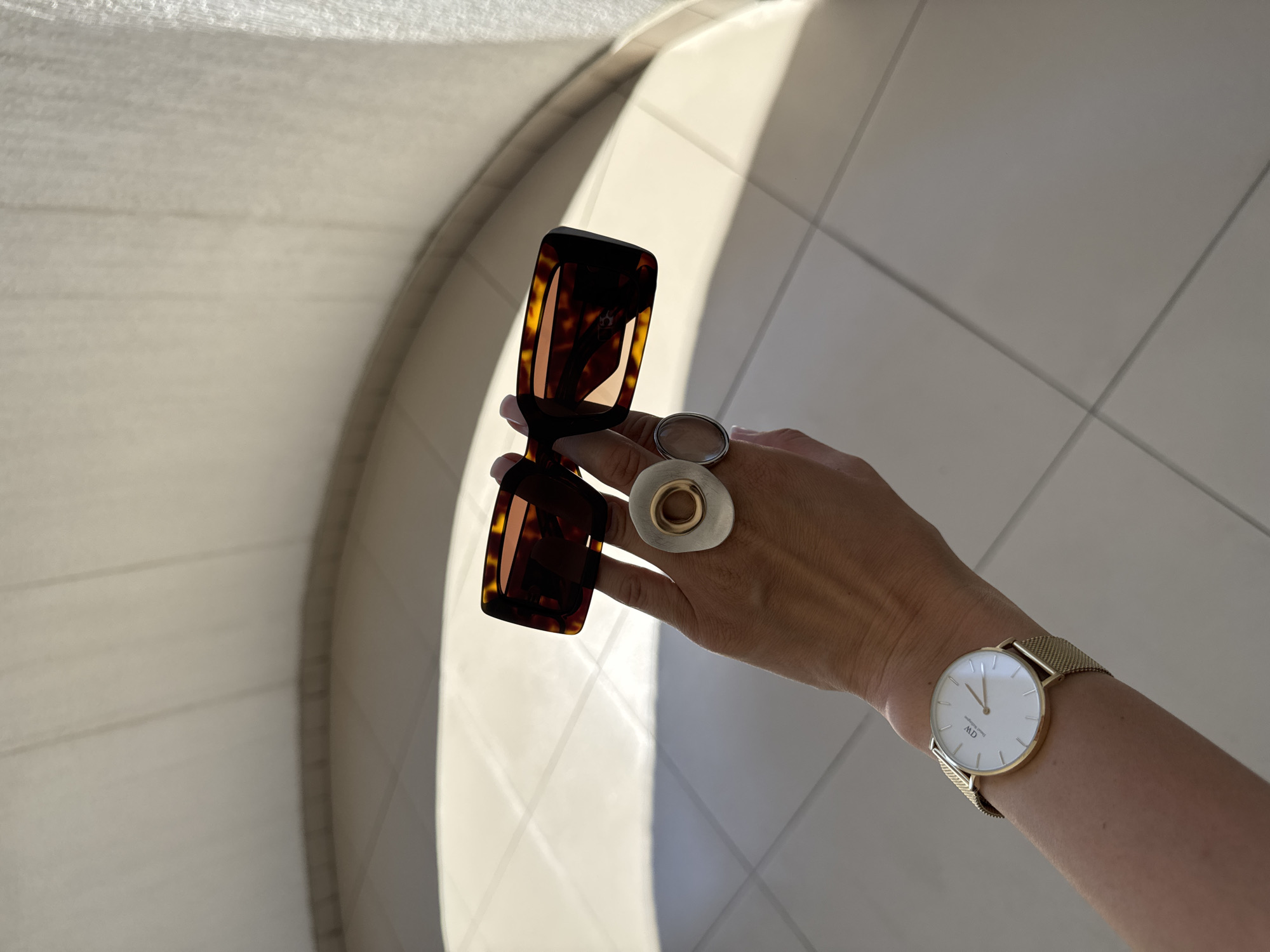 A person's hand wearing a gold mesh watch and two rings is holding a pair of tortoiseshell sunglasses. The background appears to be an indoor tiled floor with a curtain in the upper right corner. The lighting suggests it might be daytime, possibly near a window.