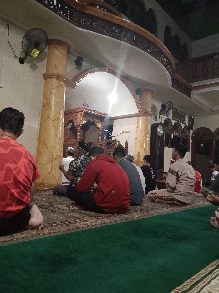 The image shows a group of men in a mosque in Tugu, Indonesia, located at the coordinates -6.976 latitude and 110.331 longitude. The men are sitting on a green carpet, facing a wooden pulpit, in the center of the room. The mosque has large pillars, a ceiling with intricate wood carvings, and a clock on the wall. The men are wearing traditional clothing and are engaged in prayer. One man is standing at the pulpit, addressing the congregation. The mosque is well-lit and the scene is quiet and serene.