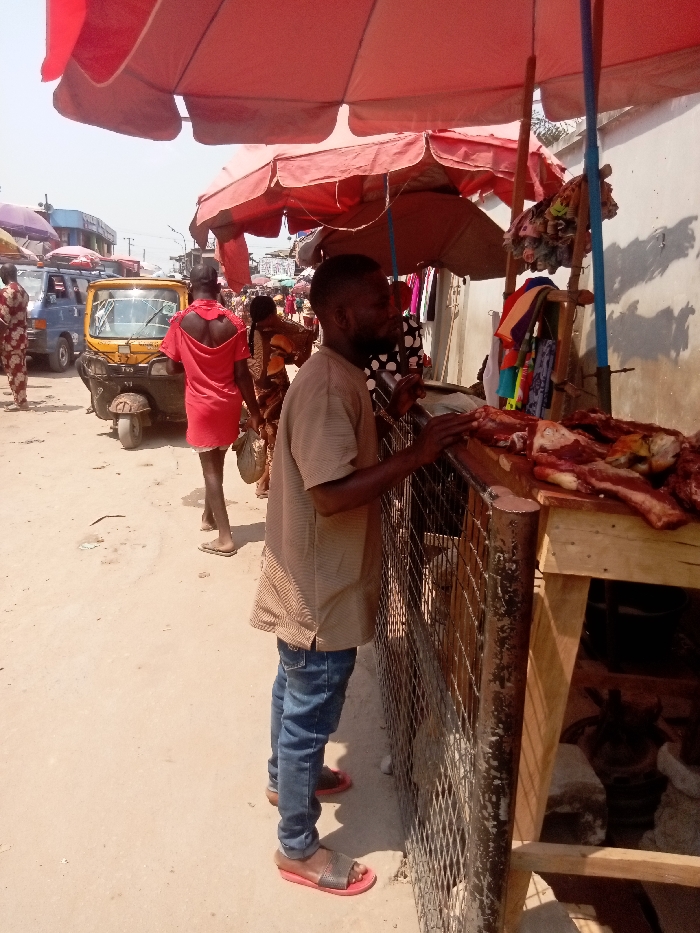 The image depicts an outdoor market scene in Ogidi, Nigeria, under daylight conditions. In the foreground right, a market stall displays raw meat cuts, including ribs and other portions, on a wooden surface. A dark-complexioned man with a short beard, wearing a light brown striped short-sleeved top, blue jeans, and red-soled sandals, stands facing the stall, leaning with his hands near the counter. Below the counter, behind a metal grate, the head of a horned animal, likely a goat or ram, is partially visible. A large red market umbrella provides shade over this area. To the left, a second dark-complexioned man in a red short-sleeved top and flip-flops walks away from the viewer. Behind him is a yellow Keke Napep (motorized tricycle taxi). A blue van is partially visible further in the background, amidst more market activity, additional vendors, and umbrellas. The ground is composed of packed dirt. A light-colored building wall forms the background on the right, exhibiting shadow patterns.