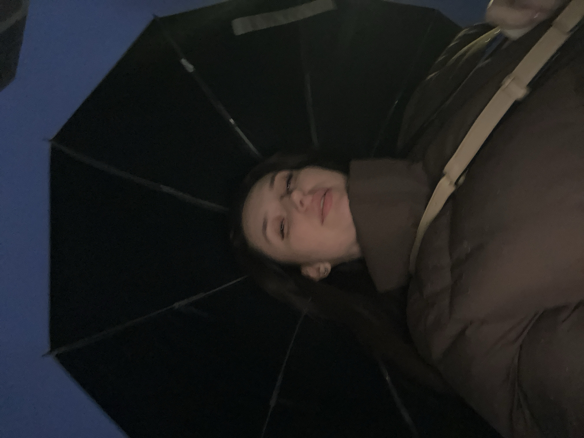 A young woman is depicted in a low-angle, possibly selfie-style, shot taken outdoors at night in Tyumen, Russia. Her face, positioned towards the right side of the frame, shows a neutral or slightly pensive expression, with her mouth slightly open. She has dark hair and is wearing a dark brown or olive-green puffy jacket, with a light-colored strap (possibly from a bag) visible across her chest. A large black umbrella, open and mostly covering the top and left side of the image, indicates protection from rain or simply its use as a prop. The dark blue background suggests the evening or night sky. No text is visible in the image.