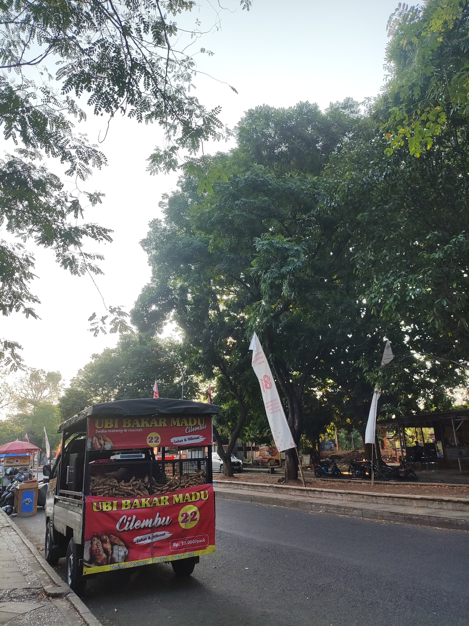This image depicts a street scene in Cirebon, Indonesia, featuring a food cart selling "Ubi Bakar Madu Cilembu," which translates to Cilembu Honey Roasted Sweet Potatoes. The cart, a modified pickup truck, is parked on the side of a paved road, with a sidewalk in the foreground. The truck is adorned with large red banners advertising the product, displaying images of roasted sweet potatoes, the price of Rp. 10,000 per pack, and a phone number. Inside the truck bed, piles of sweet potatoes are visible.
Behind the food cart, a line of motorcycles is parked under the shade of large trees. In the background, there are several small structures that appear to be open-air stalls or shelters. Two white flags are visible, one prominently displaying the number "18" and text that is partially obscured. The overall environment suggests a warm, outdoor setting, possibly during daytime, with dappled sunlight filtering through the dense foliage of the trees. The sky is bright, indicating clear weather. No people are clearly discernible in a way that allows for identification or detailed description of their actions.