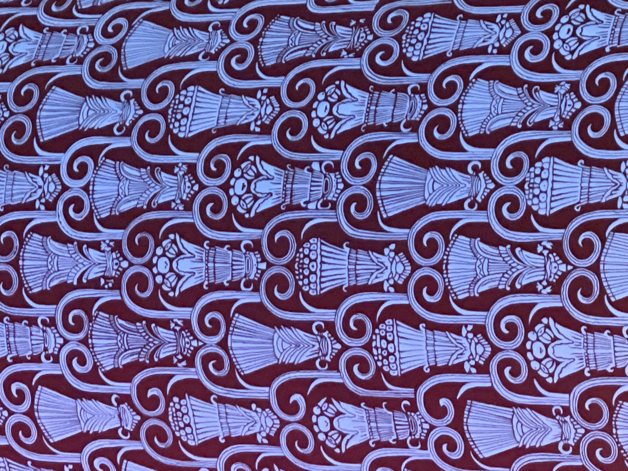 This image displays a detailed, repeating pattern on a deep red background. The pattern consists of stylized, urn-like shapes with varying floral and geometric designs. These urn shapes are arranged horizontally and vertically, connected by flowing, swirling lines, creating a continuous, decorative motif. The lighter color of the pattern elements contrasts sharply with the dark background, enhancing the visibility and complexity of the design. The overall impression is one of an artistic or decorative textile or wallpaper. It is known that this patterned textile or wallpaper is located in the city of Madrid, Spain.