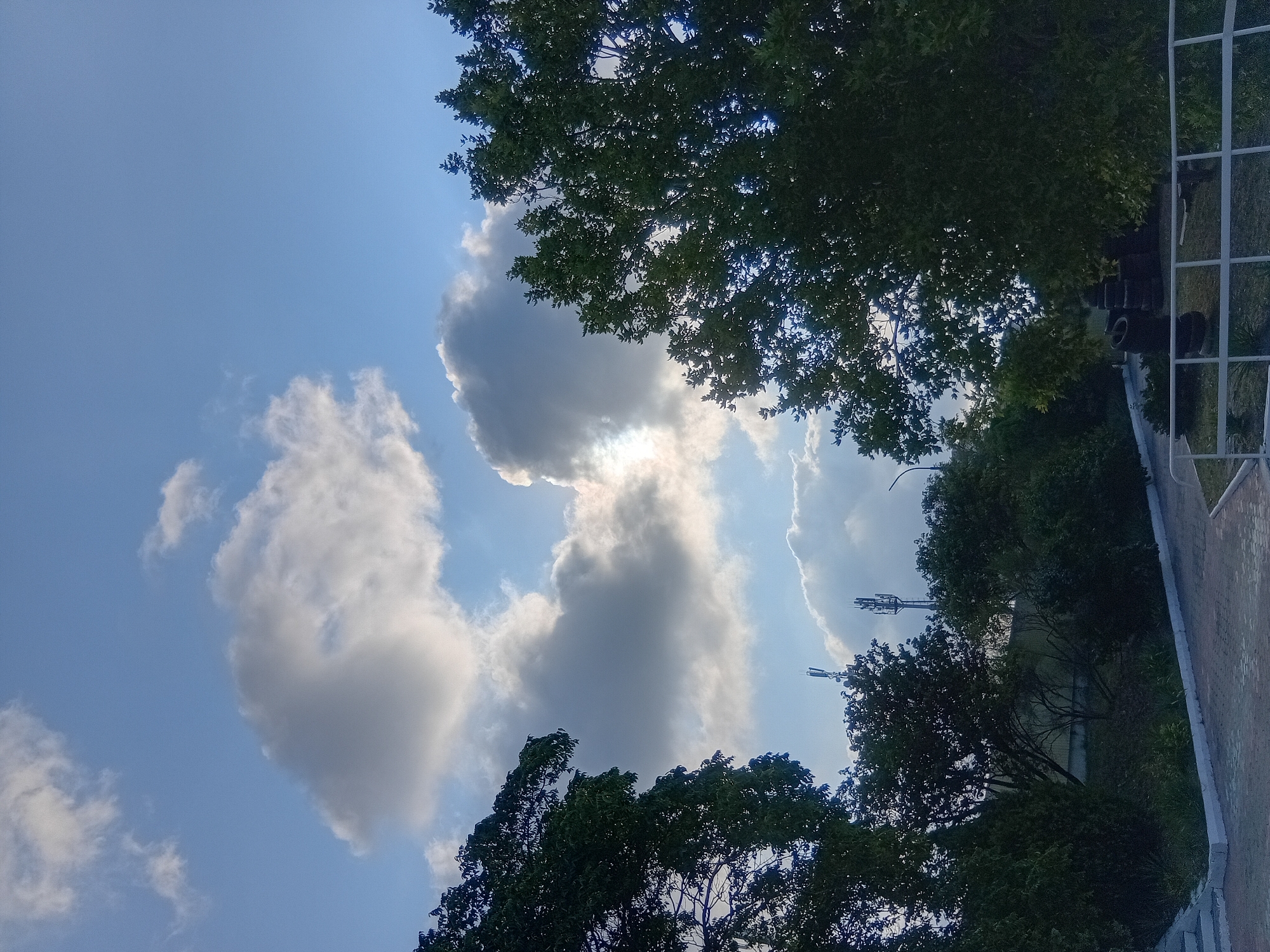 The image depicts a partially cloudy sky above a park-like area with dense foliage. Tall trees with dark green leaves frame the scene from the left and right sides. In the middle ground, there are more trees and some shrubbery. A paved pathway is visible in the foreground, bordered by a white railing and some stacked tires. Two communication towers are also visible in the background amidst the trees. The sky is a mix of blue and white, with sunlight partially obscured by clouds. The overall impression is an outdoor, possibly urban or suburban, green space during daylight hours.
