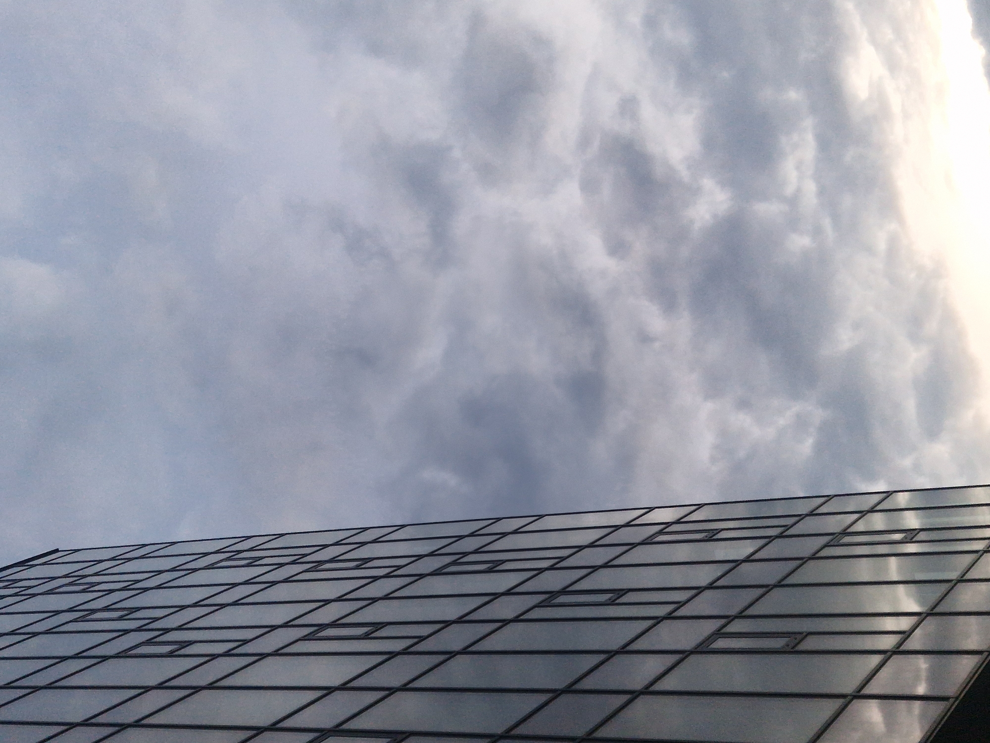 The image presents a low-angle perspective of a modern architectural structure, likely the roof or upper facade of a building, set against a cloudy sky. The structure is composed of numerous large, dark, rectangular and trapezoidal glass panels, separated by thin, dark frames. These glass panels are highly reflective, mirroring the cloudy sky above. Several smaller, rectangular openings or windows are visible within some of the larger glass sections.
The sky dominates the upper portion of the frame, filled with dense, textured clouds that vary in shade from light grey to darker, almost stormy grey. Towards the right side of the image, the clouds transition into a brighter, almost glowing white and light yellow area, suggesting the presence of strong light, possibly sunlight, breaking through or reflecting off the cloud cover.
No people or discernible activities are present in the scene. The weather is clearly cloudy, and the lighting, with its bright highlights amidst the overcast conditions, suggests a time of day when the sun is either breaking through or nearing the horizon. The architectural style appears contemporary. There is no visible text within the image.