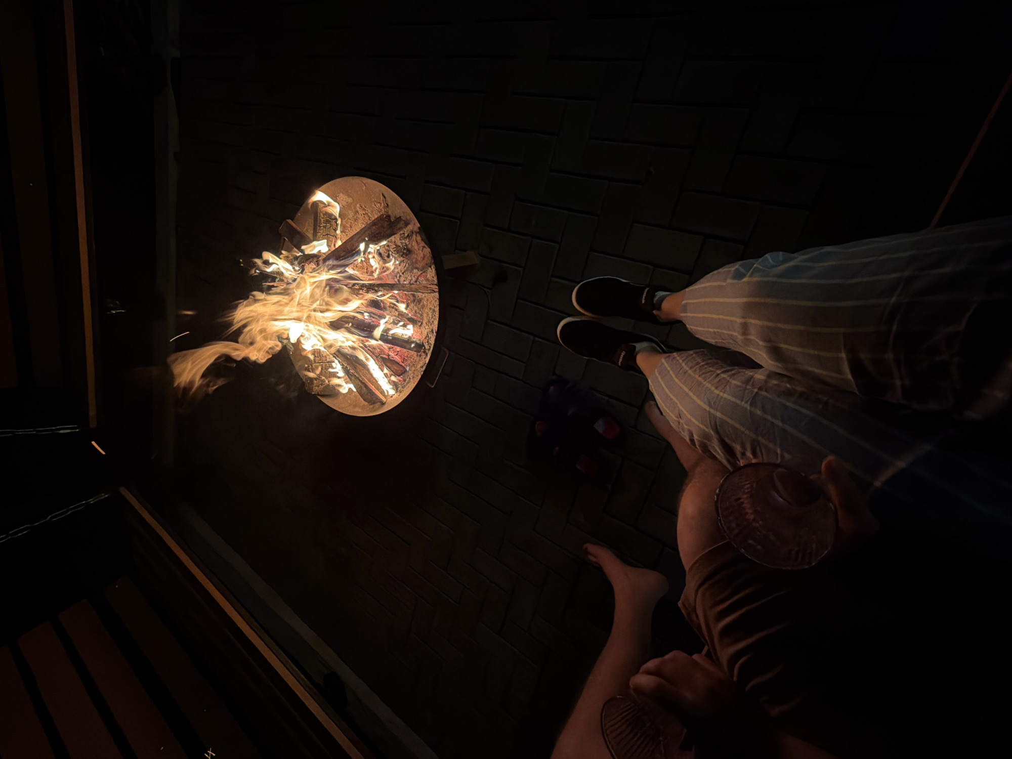 The image depicts a nighttime gathering in Kamennoye, Russia. A circular fire pit filled with burning wood is prominently featured, casting light onto a brick-paved ground. Two individuals are visible: one is wearing striped pants and black shoes, the other is barefoot and holding a glass. A dark object, possibly a bag or container, is placed near the fire pit. The surrounding environment appears to be a dark outdoor space, with a glimpse of a wooden structure on the left.