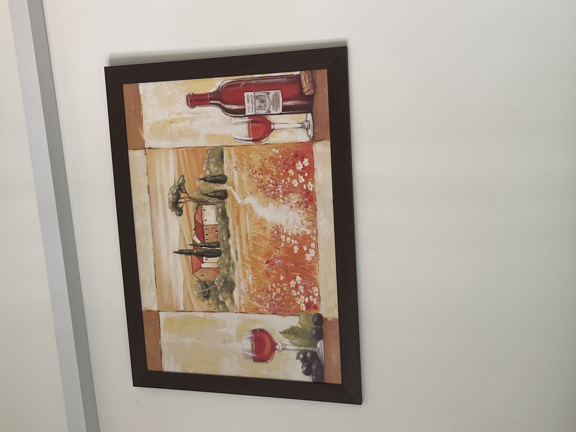 An unframed, rectangular painting depicts a Tuscan-inspired landscape. The foreground features a glass of red wine and a bunch of dark grapes on the left, and a bottle of red wine with a glass and a cork on the right. The middle ground shows a pathway winding through a field of small, scattered white and orange flowers. In the background, there are several red-roofed buildings with cypress trees. The sky is a soft orange and yellow. The painting is hung on a plain white wall, with a section of a light grey ceiling molding visible at the top of the image. The painting is oriented horizontally. The scene described is a painting and does not depict any specific location in the city of Pryluky, Ukraine.