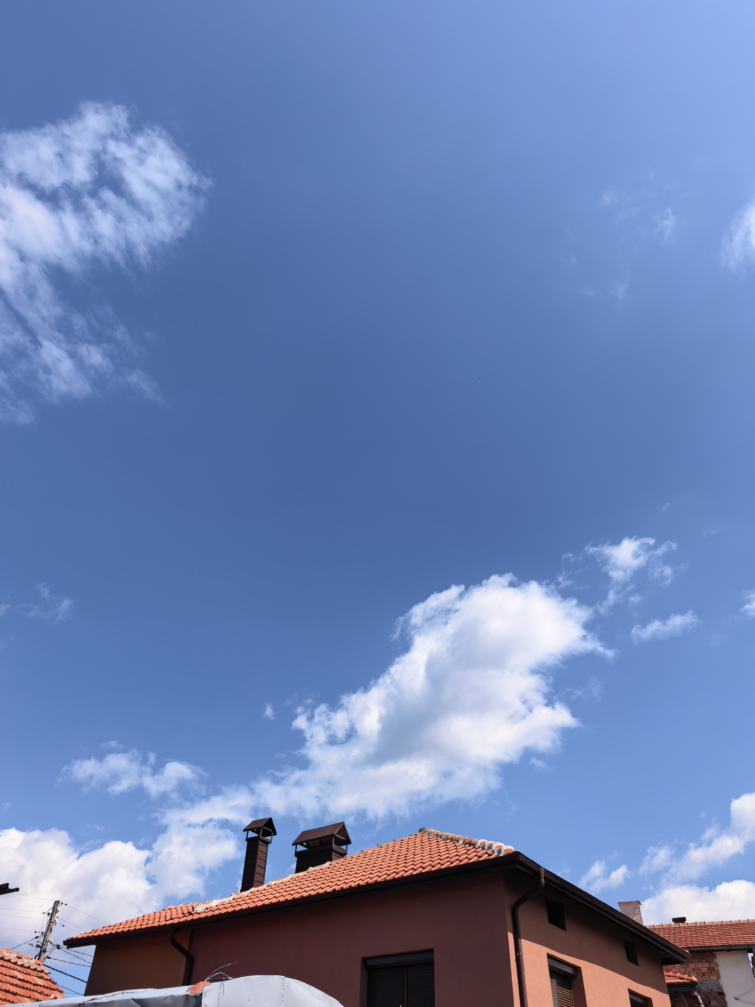 A medium-angle shot depicts a residential area in Unknown, Unknown. The sky is predominantly blue with scattered, fluffy white clouds. In the lower portion of the frame, the roofs of buildings are visible, characterized by terracotta tiles. Chimneys extend upwards from the roofs. The buildings are painted in a terracotta hue, harmonizing with the roof color. An electrical pole with wires stretches across the scene in the lower left corner. The scene conveys a sense of calm and clear weather.
