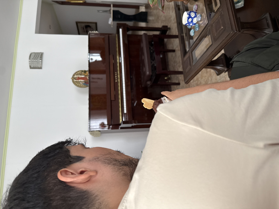The scene depicts an interior residential space within the city of AMCO, Area Metropolitana Centro Occidente, Colombia. A male individual is visible from the back and right side, wearing a white t-shirt. He has dark hair and stubble along his jawline. He holds a transparent plastic container containing a dark dessert, surmounted by a heart-shaped wafer. In the background, a brown Kawai upright piano is positioned against a white wall, accompanied by a matching brown piano bench. A golden-framed decorative object is mounted on the wall directly above the piano. To the right of the piano, an open doorway reveals an adjacent room. A wall-mounted light fixture is visible on the upper section of the white wall. In the right foreground, a dark wooden coffee table with a glass top holds a small blue and white patterned ceramic cup and multiple smooth, painted stones. A potted plant is partially visible on the table's right edge.
