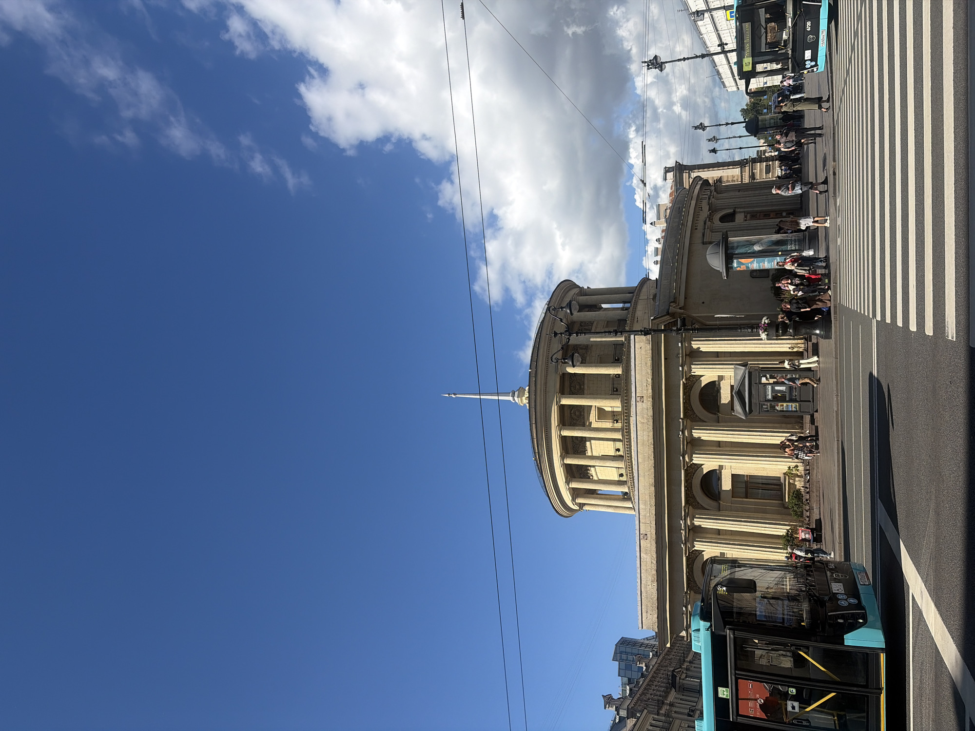 The image depicts a street scene in Saint Petersburg, Russia. The primary subjects are people, a blue bus, and a large, classically designed building with columns. The scene is set on a street with a pedestrian crossing, indicated by white stripes.
The activity taking place appears to be a normal day on a busy street. People are walking and standing on the sidewalk and near the pedestrian crossing. The bus is stopped, possibly at a bus stop, with its doors open. Some people are boarding or disembarking.
Notable details include the bright blue sky with scattered clouds, suggesting fair weather. The time of day appears to be daytime, likely late morning or afternoon, given the sunlight and shadows. The architecture of the building suggests a historical or cultural landmark. The prominent spire on the building's dome is a distinctive feature.
Visible text includes some Cyrillic lettering on the building, appearing to be "МЕТРОПОЛЬ" (METROPOL) above a doorway. There is also text on the bus, including the number "4720" and Cyrillic characters. Advertising posters are visible on display cases near the building entrance. The overall mood of the scene is one of everyday urban life.