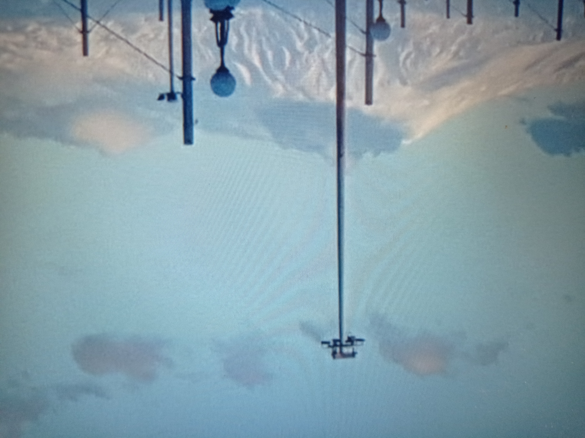 The image depicts a wide shot of a snow-covered mountain range under a cloudy, pale blue sky. In the foreground, several tall poles are visible, some with lights or other attachments. One prominent pole in the center has a cross-shaped fixture at its top, suggesting it's a lighting or signaling tower. Another pole to the right has a round, globe-like fixture. The mountain landscape is textured with shadows and highlights, indicating uneven terrain and possibly volcanic formations. The lighting suggests it could be early morning or late afternoon. The scene is devoid of people. The image does not provide any specific landmarks or context to confirm its location in Mukachevo, Ukraine.