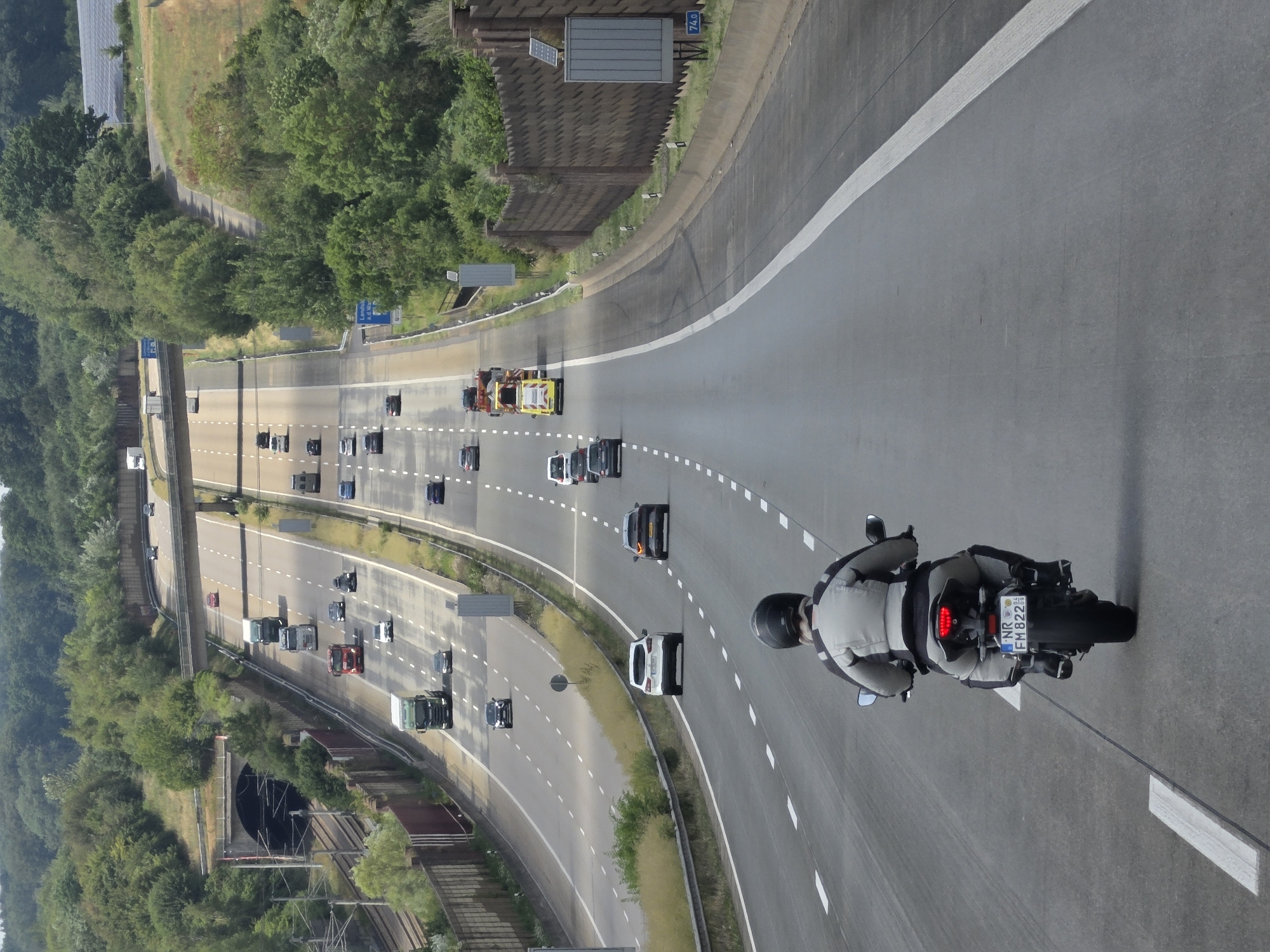 An aerial, long shot captures a multi-lane highway in Oberhaid, Germany, with a motorcycle rider in the foreground heading away from the viewer. The motorcycle, a dark color with a license plate reading "NR EM 822", is in the right lane. The rider is wearing a light-colored motorcycle jacket and black pants with a black helmet. Numerous cars and trucks are visible on the highway, traveling in both directions. To the left of the highway, a train tunnel entrance is visible, with train tracks running alongside it. The highway is surrounded by lush green trees and vegetation, with a concrete barrier along the right side of the road. The sky is overcast.