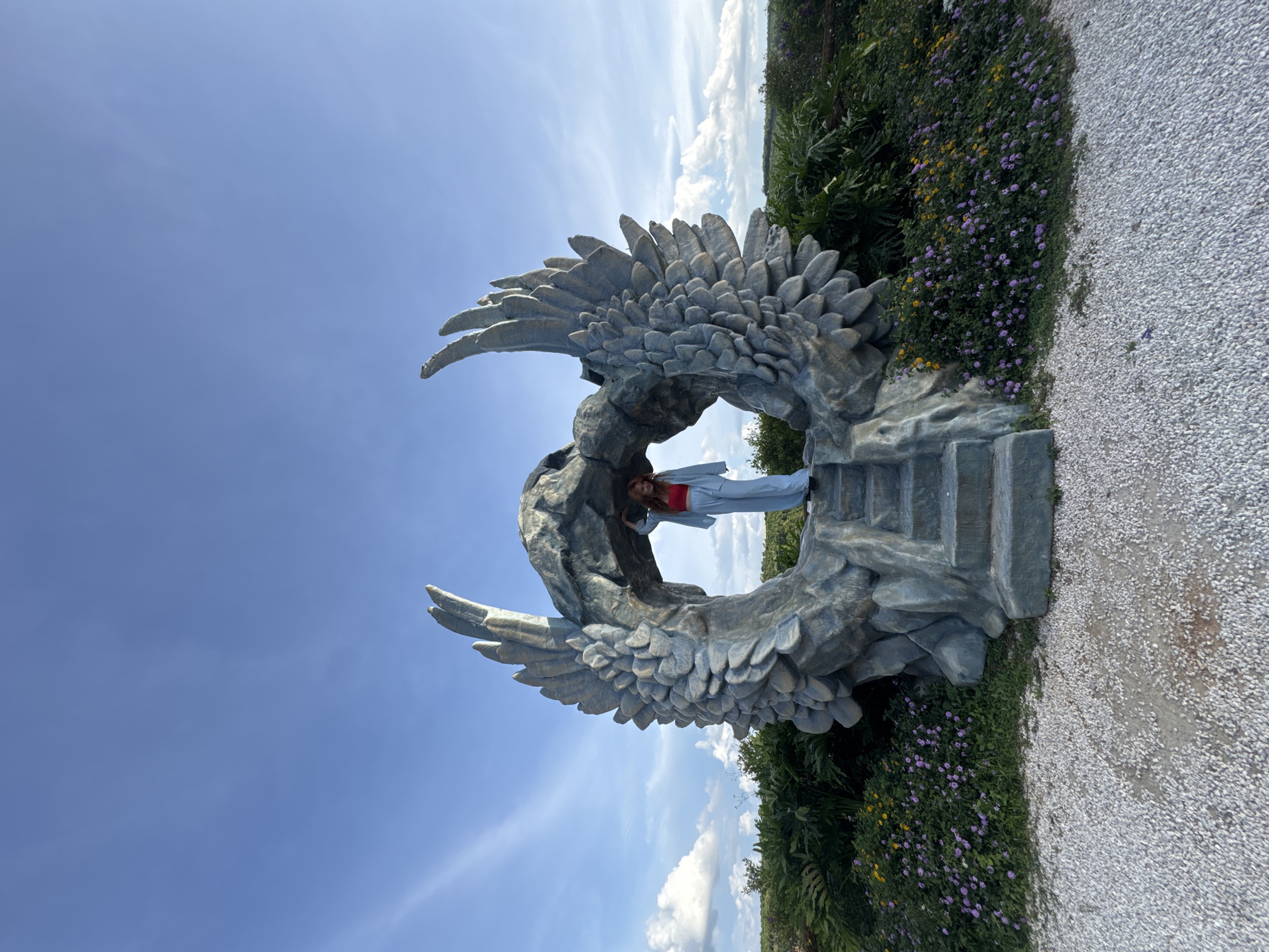 A woman stands on a stone staircase leading into a large, gray, rock sculpture resembling a pair of angel wings arching over a cave-like opening. She wears a red top and light blue pantsuit. The sculpture is surrounded by lush greenery and small purple and yellow flowers. In the background, a vast expanse of blue sky with scattered white clouds stretches above a landscape of rolling hills. The scene appears to be outdoors in a park or garden setting, likely in Thôn 1, Vietnam, given the context of the request. The ground in the foreground is covered in white gravel.