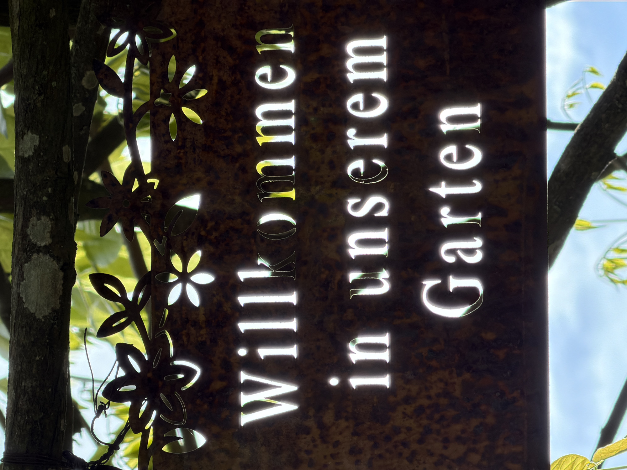 A vertical garden sign in German, "Willkommen in unserem Garten," (Welcome to our Garden) hangs from a tree. The sign is rusted metal with flower-shaped cutouts on its left side. The words are cut out of the metal to allow light to shine through. The sign is set against a natural background of trees and sky. This is located in the city of Esch-sur-Alzette, Luxembourg.