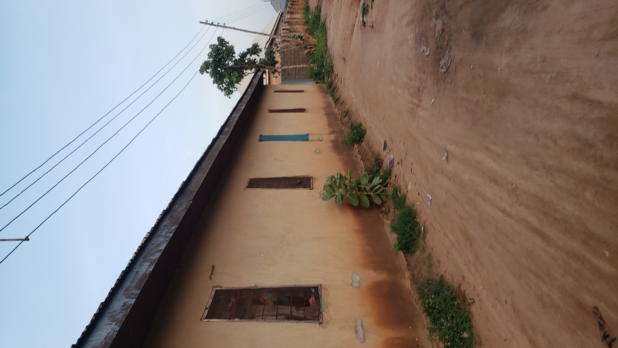The image depicts a street scene in Jauro Yinu, Nigeria, characterized by a long, light-colored building running along the left side of a dirt road. The building has a corrugated metal roof and several windows, some of which are covered with metal bars. A plant with large, succulent leaves grows near the building, and other sparse vegetation is visible along the side of the road, which is littered with small pieces of debris. Overhead, power lines crisscross the sky, supported by a utility pole visible in the distance. The overall environment suggests a residential or commercial area in a developing region, with simple construction and a natural, unpaved road surface.