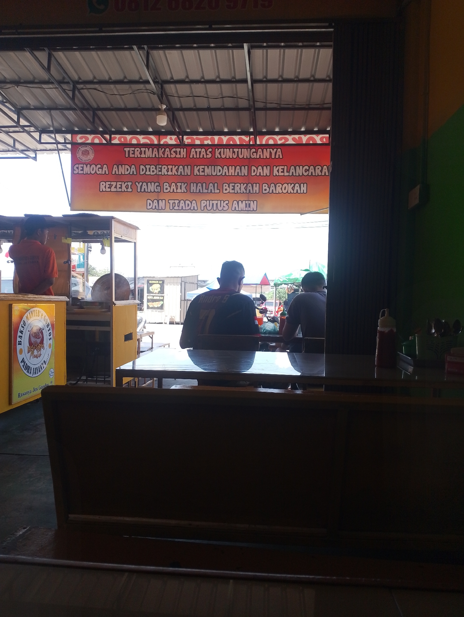 An outdoor dining area in Batam, Indonesia, appears to be a food stall or restaurant. In the background, a red and orange banner reads "TERIMAKASIH ATAS KUNJUNGANYA SEMOGA ANDA DIBERIKAN KEMUDAHAN DAN KELANCARA REZEKI YANG BAIK HALAL BEROKAH DAN TIADA PUTUS AMIN." This translates to "THANK YOU FOR YOUR VISIT, MAY YOU BE GRANTED EASE AND SMOOTHNESS, GOOD HALAL BLESSINGS, AND UNINTERRUPTED WEALTH, AMEN." A person in an orange shirt stands behind a food cart. Two men sit at a table, one wearing a dark shirt with a number 71 on the back. A bottle of sauce and cutlery are visible on a table in the foreground. The scene is illuminated by natural daylight.