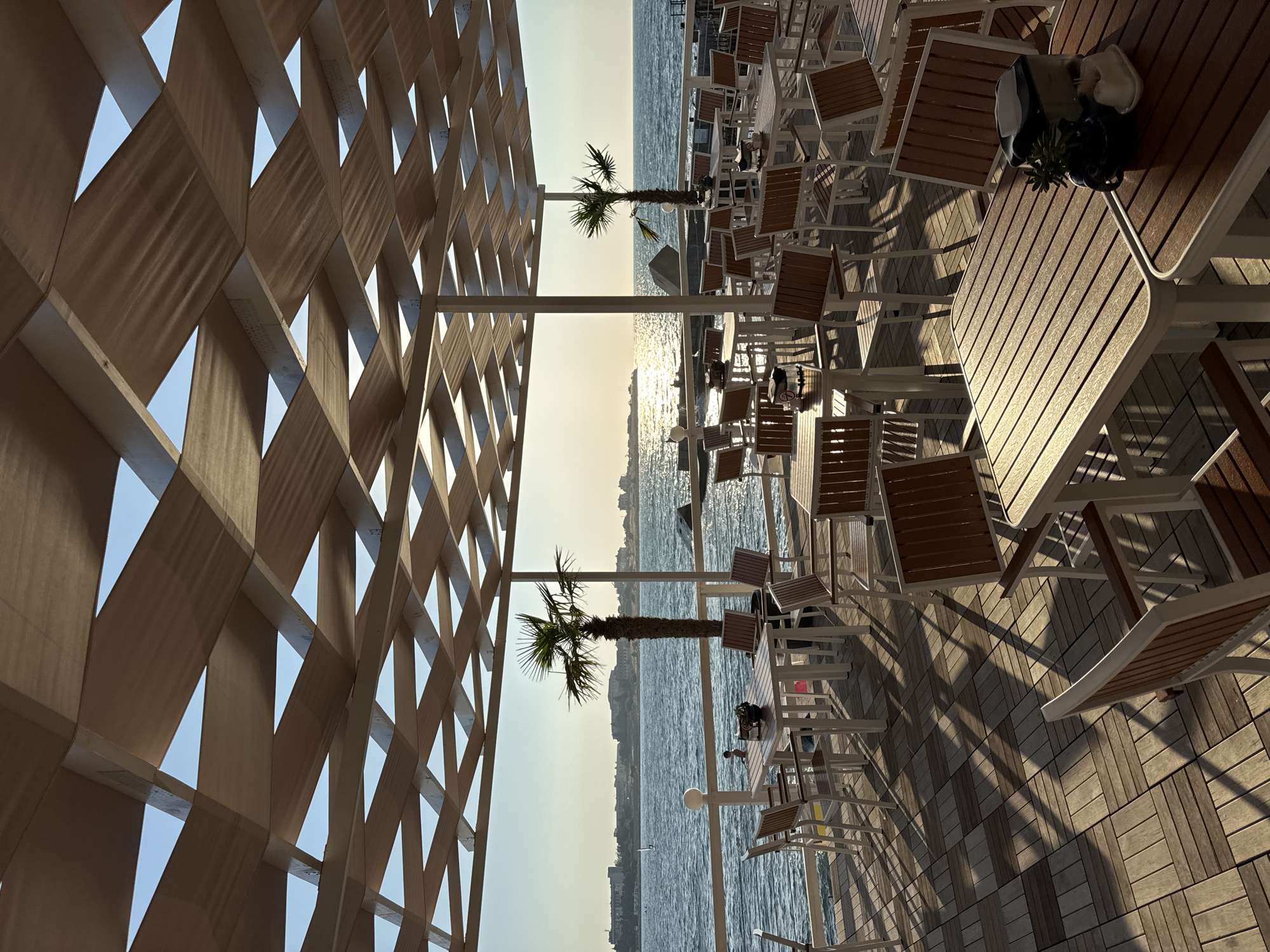 This image depicts an outdoor seating area of a restaurant or cafe located in Sevastopol, Ukraine, facing the sea. The area is covered by a distinctive lattice-like wooden canopy that casts geometric shadows on the wooden deck below. Numerous tables and chairs, made of wood and white metal, are arranged on the deck.
In the background, the sea stretches out to the horizon, with a hazy cityscape visible along the coastline. The sun is setting or rising, casting a warm glow on the water, and its reflection is prominent. Two palm trees are present, one taller and more prominent than the other, adding to the seaside ambiance. A few people are visible in the distance, including one individual near the water's edge and a sailboat on the sea. The overall scene suggests a peaceful, open-air dining environment by the Black Sea.
