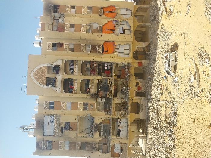 An image taken in Khan Yunis, Palestinian Territory, depicts a heavily damaged multi-story residential building under a clear blue sky. The building's light-colored facade is riddled with numerous small impact holes, consistent with bullet strikes, and shows extensive structural damage, including blown-out sections of walls and windows on multiple floors. Exposed interiors reveal rubble and debris. Dark burn marks are visible on the upper right side of the structure. White water tanks and communication equipment are situated on the roof. Balconies on the upper-middle floors show remnants of clothes hanging. The ground immediately in front of the building is a vast expanse of sand and severe rubble, including shattered concrete blocks and twisted metal. In the mid-ground, a heavily damaged, inverted white vehicle chassis is visible amidst the debris. A solitary, indistinct figure stands within the shadowed arcade area of the building's ground floor on the right.