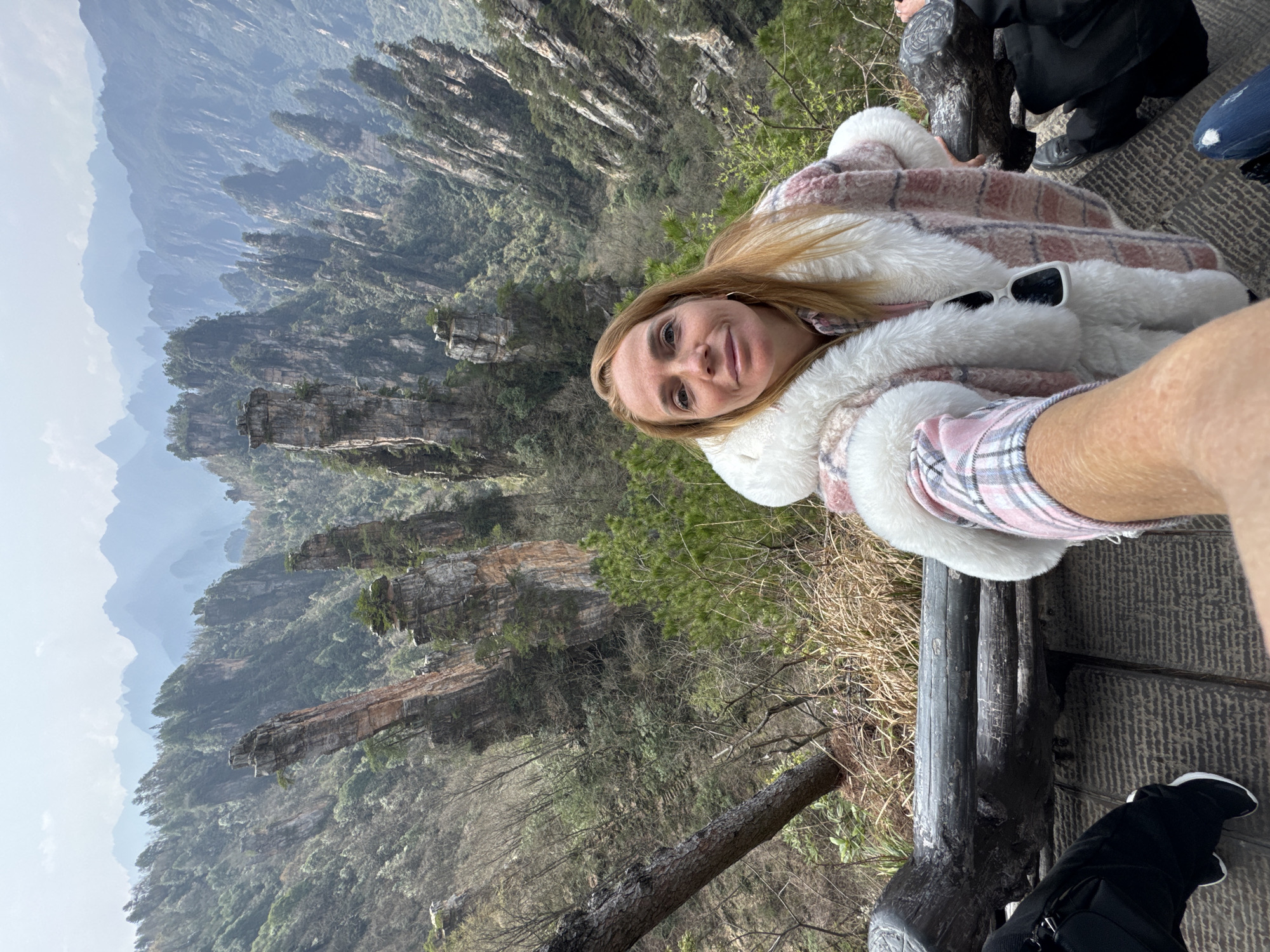 A selfie is taken from a wooden viewing platform overlooking a deep valley filled with numerous tall, verdant sandstone pillars and peaks in Wulingyuan, China. An adult female, appearing Caucasian with long light brown hair and a faint smile, occupies the right foreground. She wears a thick, pink and white plaid coat with extensive white faux fur trim, and white sunglasses are tucked into her collar. Her left hand is partially visible, suggesting she is either holding the camera or being photographed by the person whose bare, freckled forearm extends from the lower left towards her. The wooden platform, visible in the lower frame, supports a person wearing black pants and black and white sneakers on the left, and another in blue jeans and black shoes on the far right. A dark, weathered wooden log railing separates the platform from the steep, densely forested drop-off. Beyond the railing, the landscape features dramatic, tree-covered sandstone formations, typical of the Zhangjiajie area, receding into a hazy, blue-tinted distance under an overcast sky.