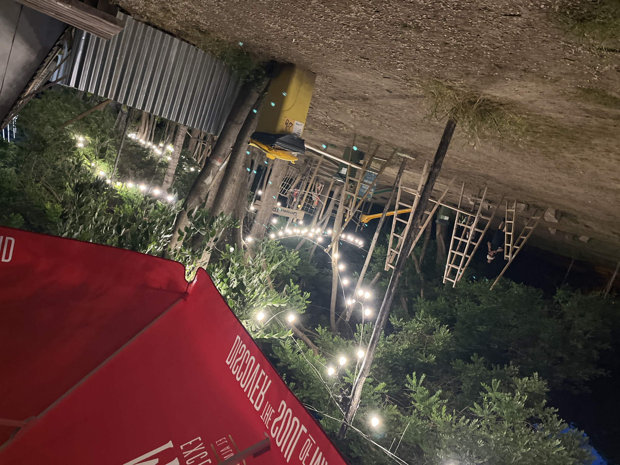 This image depicts an outdoor area at night, likely a park or recreational space. The scene is illuminated by string lights draped between trees. In the foreground, a large red umbrella is partially visible, displaying white lettering that is partially obscured. To the left, a person is standing near a wooden ladder. A yellow trash bin with a black bag and a yellow object on top is located in the middle ground. In the background, more wooden ladders and trees are visible, along with what appears to be a climbing structure. A corrugated metal fence runs along the right side of the image. The ground is composed of dirt and gravel. The overall impression is of a casual, outdoor gathering or event. It is not possible to determine the specific location in Plovdiv, Bulgaria from the provided image.