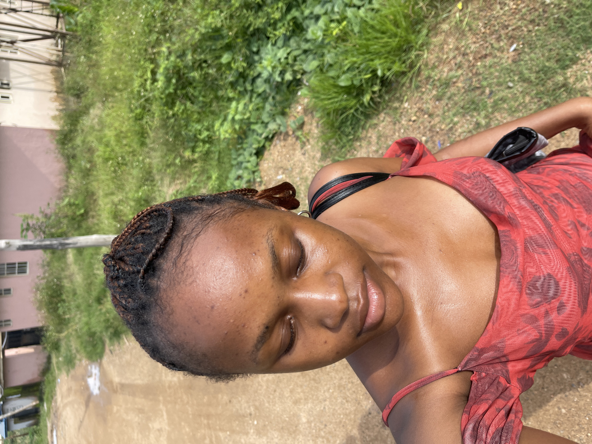 A high-angle, medium shot captures a Black woman looking down with her eyes closed, standing outdoors on a dirt path. She has braided hair and is wearing a red off-the-shoulder dress. A black purse is slung over her right shoulder. The background consists of tall green grass, a pale pink building, and other structures suggesting a residential area. The image appears to have been taken in Rafin Par, Nigeria.