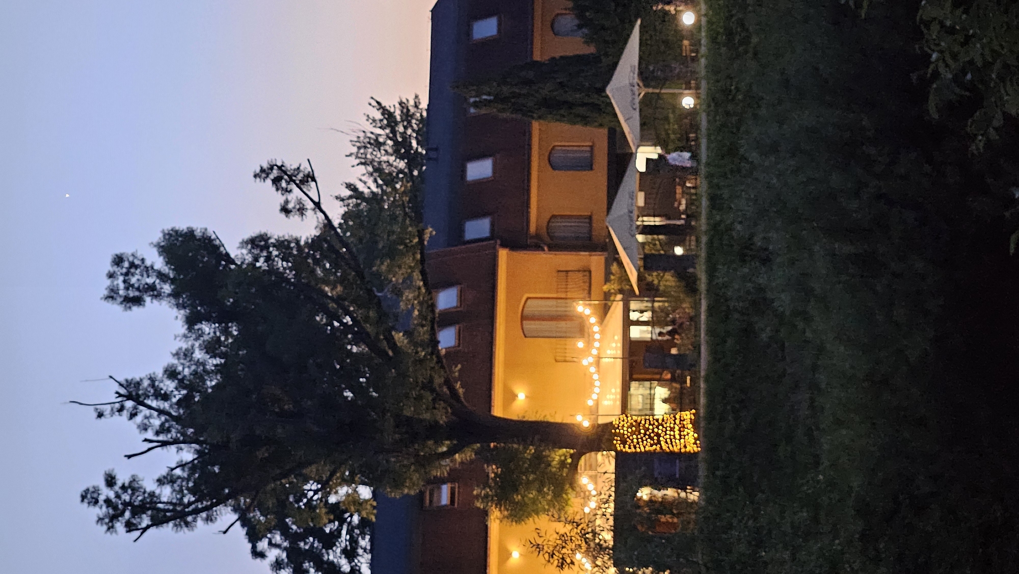 This media file captures an outdoor scene at dusk, likely in Prešov, Slovakia. The primary subjects are buildings with a warm yellow facade, adorned with strings of glowing lights. A large, mature tree with dark green foliage dominates the left side of the frame, its branches reaching towards the sky. The sky is a soft gradient of pale blue and pink, indicating the transition from day to night.
Several windows are visible on the buildings, some illuminated from within. Outdoor seating areas are visible with tables and chairs, suggesting a restaurant or cafe setting. White umbrellas are set up over some of these tables. The ground is covered in lush green grass, creating a contrast with the dark foliage of the tree and the illuminated buildings.
The activity taking place appears to be a quiet evening ambiance, possibly with patrons enjoying the outdoor space. The string lights create a festive and inviting atmosphere. The time of day is clearly evening, as evidenced by the twilight sky and the artificial lighting. The weather seems calm, with no indication of rain or wind.
Notable details include the crescent moon visible in the upper left portion of the sky. The architecture of the buildings, with their arched windows and simple, yet warm design, contributes to the overall feel of the scene. The image evokes a sense of relaxation and peacefulness, as the day concludes and the evening lights begin to twinkle. No visible text is present in the image.