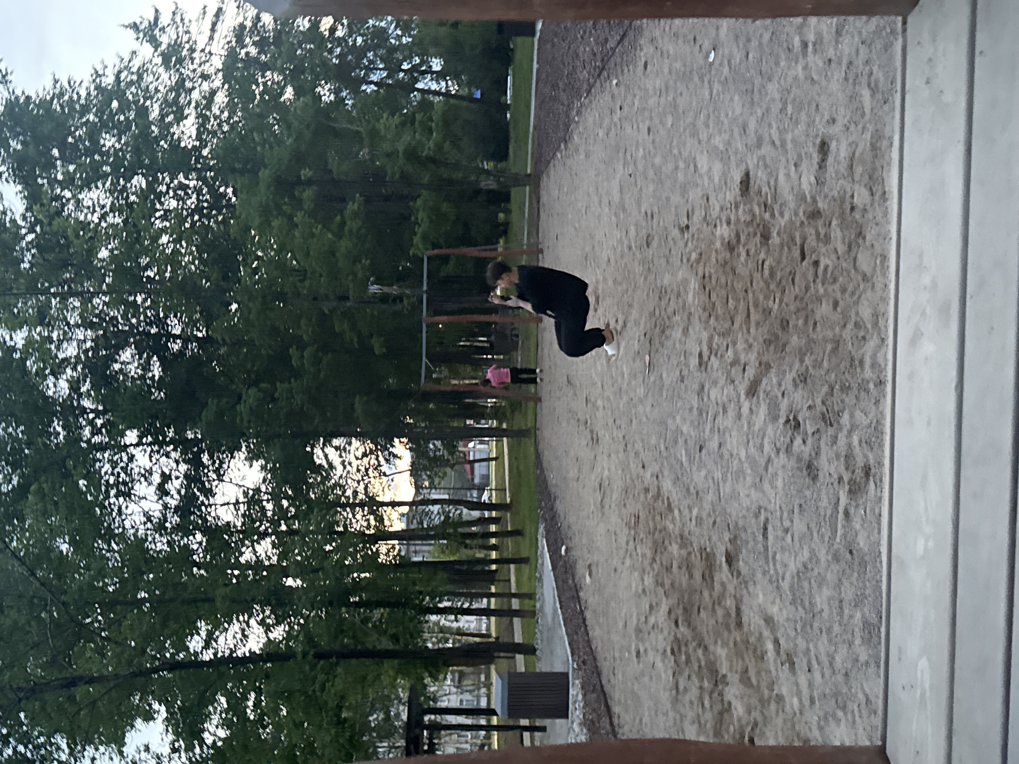 The image captures an outdoor scene, likely a park or recreational area, in Birobidzhan, Russia. The primary subject is a person, positioned in the middle ground towards the right, seen in profile. They are wearing dark clothing (black long-sleeved top and pants) and white shoes, and are in a deep crouch or lunge, looking down at something held in their hands, possibly a mobile phone or a small object.
To the left and slightly behind this main subject, another person is partially visible, wearing a pink top. The ground in the foreground and extending to the right is a large expanse of sandy or light-colored gravelly terrain. A narrow paved path runs horizontally across the middle ground, separating this gravelly area from a strip of green grass and dense trees beyond.
The left side of the frame and the background are dominated by numerous tall, slender trees, appearing coniferous, with significant foliage. Through the trees, glimpses of distant buildings or structures can be seen. Some playground equipment, such as swings or bars, is faintly visible amongst the trees. A dark, rectangular object, possibly a trash receptacle, sits near the paved path in the lower-left corner.
The sky appears overcast or partly cloudy, suggesting soft light, possibly late afternoon or early evening. The image has a calm, casual atmosphere, depicting people engaged in leisure activities in a public outdoor space. No visible text is present in the image. The photo appears to have been taken from an elevated position or through an opening, indicated by wooden beams in the extreme foreground.