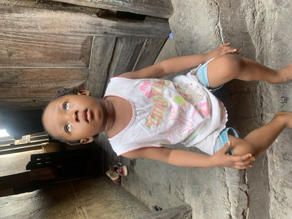 An image shows a young child seated on a concrete step in Okene, Nigeria. The child, with dark skin and braided hair, is looking upwards, with their eyes rolled back to reveal prominent sclera and their mouth slightly open. They are wearing a white sleeveless top with pink and green graphic designs and light blue shorts; they are barefoot. To the child's left, a dark, narrow passage extends into the background, where a pair of red and white sneakers and a metal bowl are visible on the ground. To the child's right, a weathered, vertically planked wooden door stands partially visible. The immediate environment consists of rough, possibly distressed concrete surfaces and aged wooden structures.
