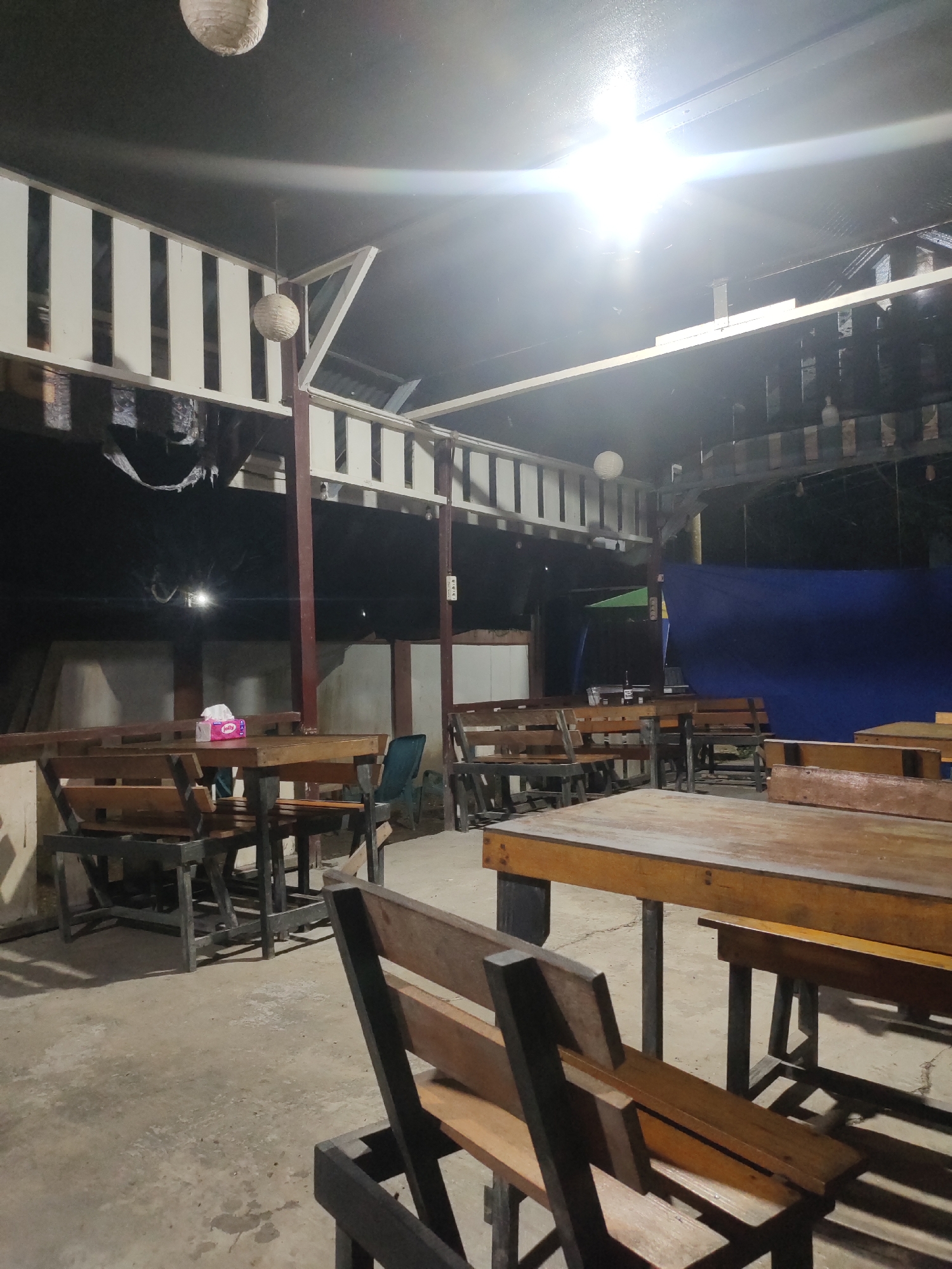 This is a photograph of an outdoor seating area at night, likely a restaurant or cafe, in Keudematangkuli, Indonesia. The scene is illuminated by artificial lights, creating shadows and a slightly dim atmosphere. In the foreground, several wooden tables and benches are arranged on a concrete floor. The tables are simple, with wooden tops and dark metal legs. Some of the benches are also visible, with wooden slats.
In the middle ground, more tables and benches are visible, some with a darker wood tone. A tissue box is placed on one of the tables. In the background, a large blue tarp hangs on the right side, suggesting a temporary or enclosed space. Above, the roof structure is visible, with white slatted railings and dark ceiling panels. Two round, woven light fixtures hang from the ceiling. The overall impression is a casual, open-air dining or gathering space. There are no people visible in the photograph.