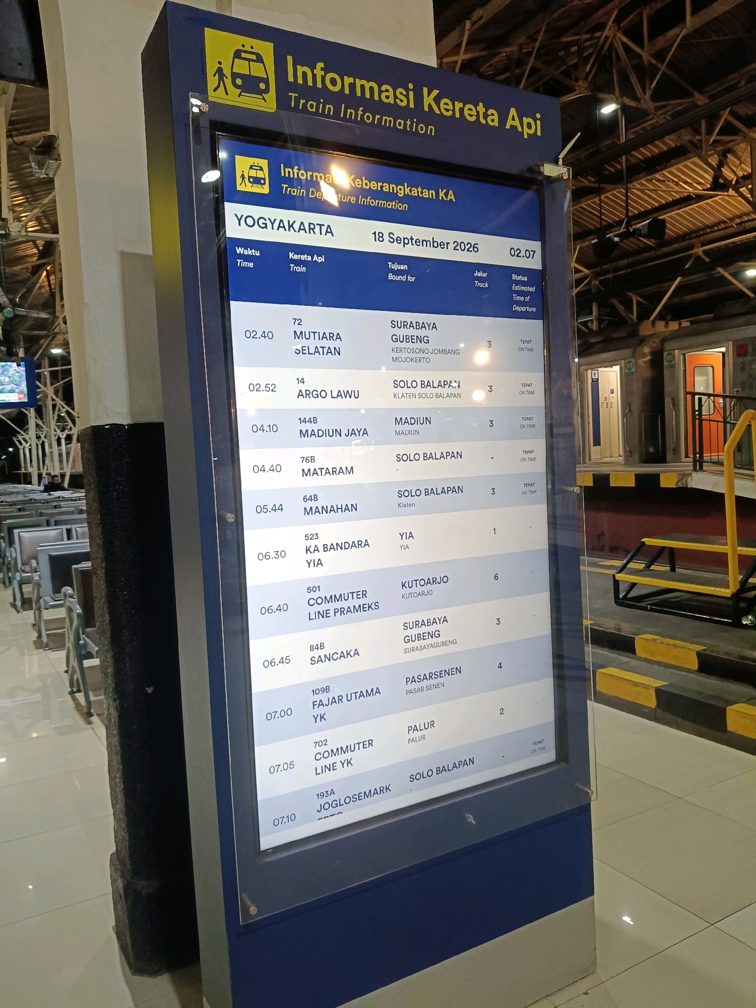 This is a medium shot, eye-level photograph of a train departure information display screen at the Yogyakarta train station in Indonesia. The screen is large and rectangular, displaying a list of trains with their departure times, destinations, tracks, and status. The screen is mounted on a dark blue stand. The background shows the interior of a train station with seating arrangements and the side of a train with orange doors. The overall lighting is dim, with the screen being the brightest element.
The screen's header reads "Informasi Keberangkatan KA" which translates to "Train Departure Information". Below this, the location is identified as "YOGYAKARTA" and the date is "18 September 2026". The columns are labeled "Waktu" (Time), "Kereta Api" (Train), "Tujuan" (Bound for), "Jalur" (Track), and "Status" (Status).
Specific train information visible includes:
- Train 72, Mutiara Selatan, departing at 02:40, bound for Surabaya Gubeng, on track 5, with a status of "Tepat On Time" (On Time).
- Train 14, Argo Lawu, departing at 02:52, bound for Solo Balapan, on track 3, with a status of "Tepat On Time".
- Train 144B, Madiun Jaya, departing at 04:10, bound for Madiun, on track 3, with a status of "Tepat On Time".
- Train 76B, Mataram, departing at 04:40, bound for Solo Balapan, on track 3, with a status of "Tepat On Time".
- Train 64B, Manahan, departing at 05:44, bound for Solo Balapan, on track 3, with a status of "Tepat On Time".
- Train 523, KA Bandara YIA, departing at 06:30, bound for YIA, on track 1.
- Train 501, Commuter Line Premeks, departing at 06:40, bound for Kutoarjo, on track 6.
- Train 84B, Sancaka, departing at 06:45, bound for Surabaya Gubeng, on track 3.
- Train 109B, Fajar Utama YK, departing at 07:00, bound for Pasarsenen, on track 4.
- Train 702, Commuter Line YK, departing at 07:05, bound for Palur, on track 2.
- Train 193A, Joglosemark, departing at 07:10, bound for Solo Balapan.
The overall scene depicts a functional information system within a public transportation hub.