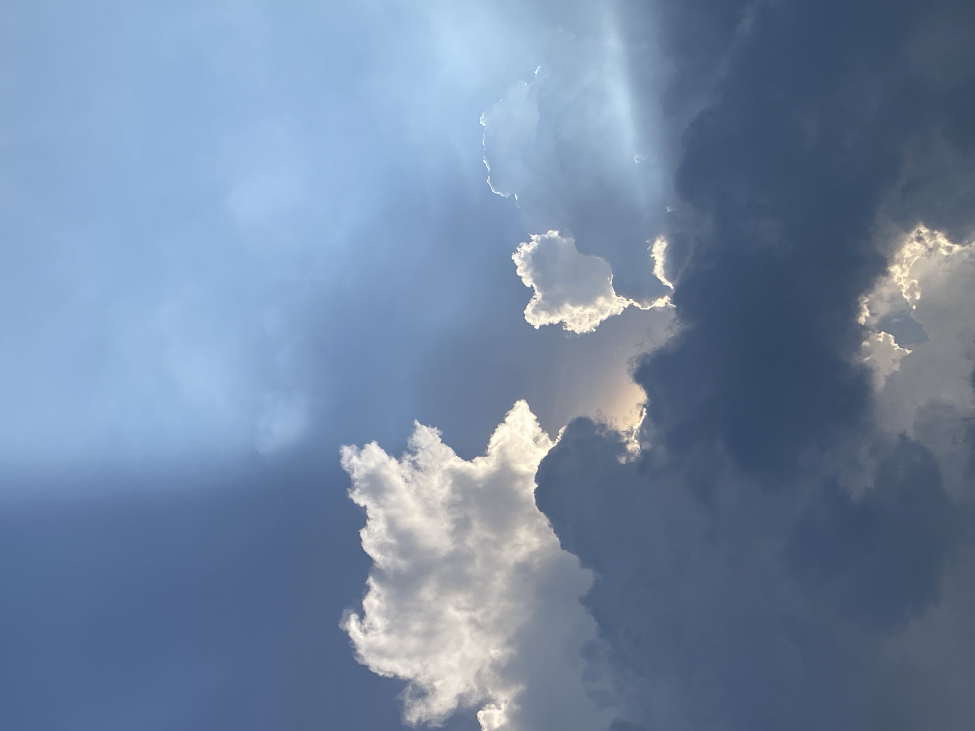 The image shows a sky with scattered clouds, some bright white and others a darker blue-grey. Sunlight appears to be streaming through gaps in the clouds, creating light shafts. The image does not depict Ho Chi Minh City, Vietnam, or any city; it only shows clouds and sky.