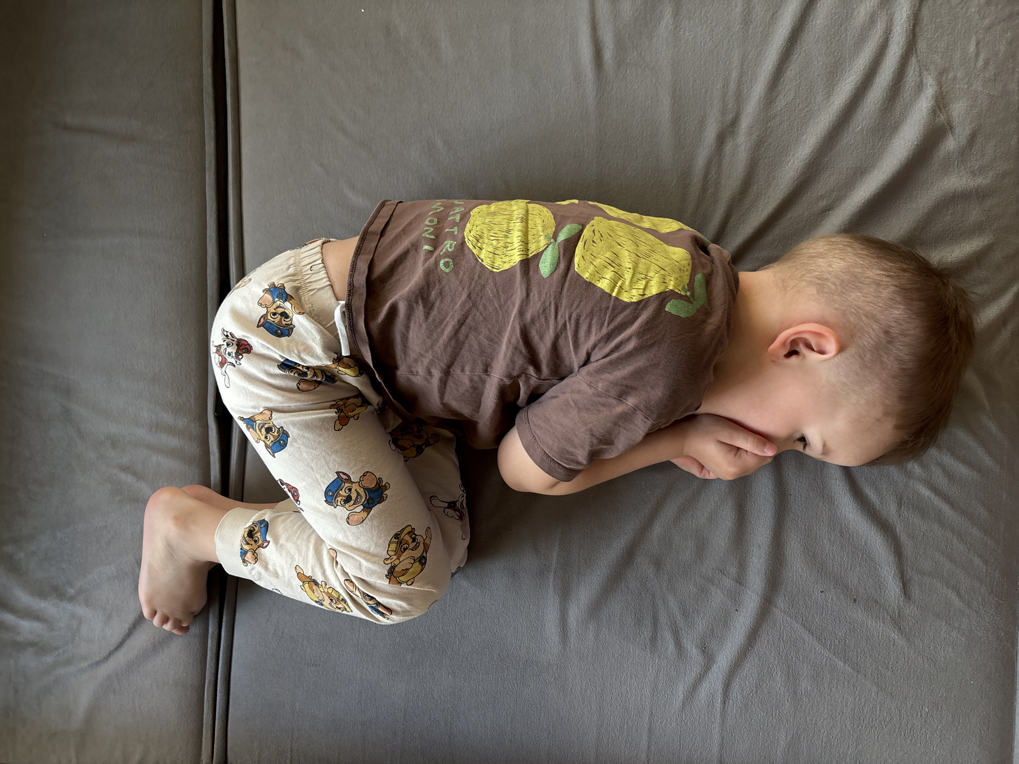 A young child, presumably a boy, is lying on a gray sofa. He is wearing Paw Patrol pajamas and a brown t-shirt with a lemon print. He is curled in a fetal position with his face buried in his hands. The location is not explicitly defined by this image, but it could be somewhere in Chaikovsky, Russia.