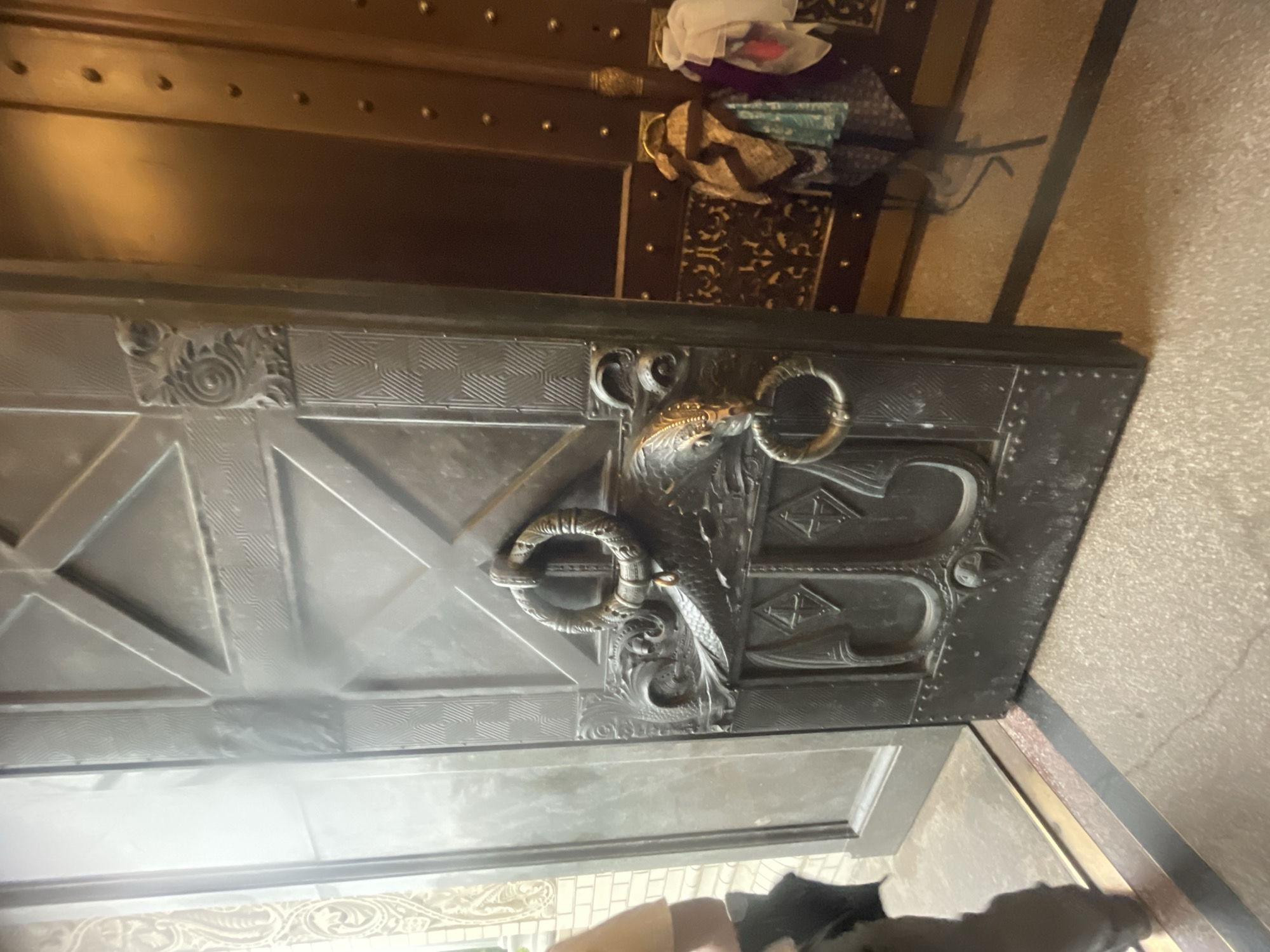The image shows a close-up of a large, ornate metal door with intricate carvings. The door is partially open, revealing another room or hallway with a wooden door and decorative trim. On the left side of the main door, a person's legs and lower torso are visible, dressed in dark pants. The floor appears to be made of stone or tile with a metallic strip. The overall scene suggests an interior space, possibly an entrance to a building of historical or architectural significance. The door features a large circular handle with a detailed design of what appears to be a dragon or mythical creature. The door itself is divided into geometric panels, with textured patterns adorning some sections. The environment appears to be dimly lit, with light coming from the left side of the image. The background on the right shows a dark wooden surface with decorative studs, and a rack with several pieces of fabric draped over it. The ornate details on the door and the overall grand impression suggest this could be a location in Kronstadt, Russia, known for its historical naval and architectural sites.
